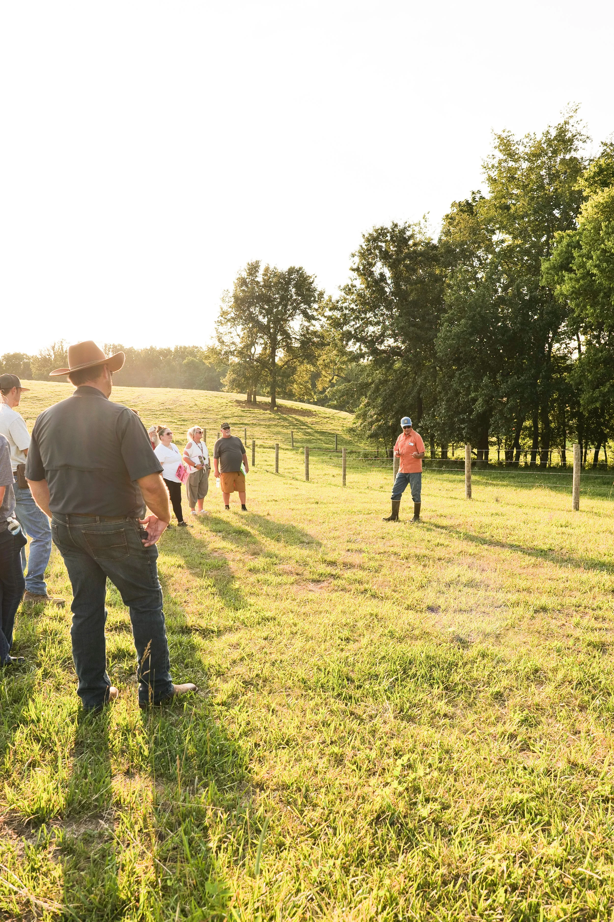 Photo by Ben Auilar. A man with an orange shirt and a ball cap stands in front of a fence line in the middle of the image. He is facing a gathering of people, about 7 of them are visible. Close to the camera is a man wearing a cowboy hat. Long shadow