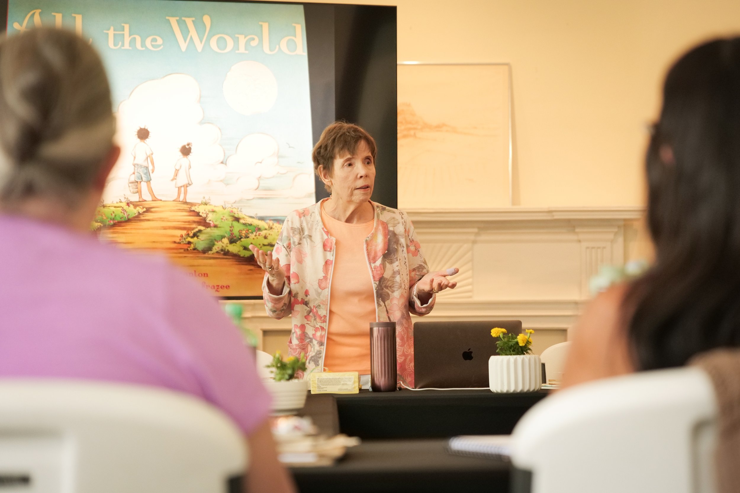Photo by Ben Aguilar. Kentucky author George Ella Lyon wears a peach colored shirt and a peach and cream flowered cardigan. She is standing in front of  a black table-clothed table; two people, whose backs are to the camera, face her. She is talking,