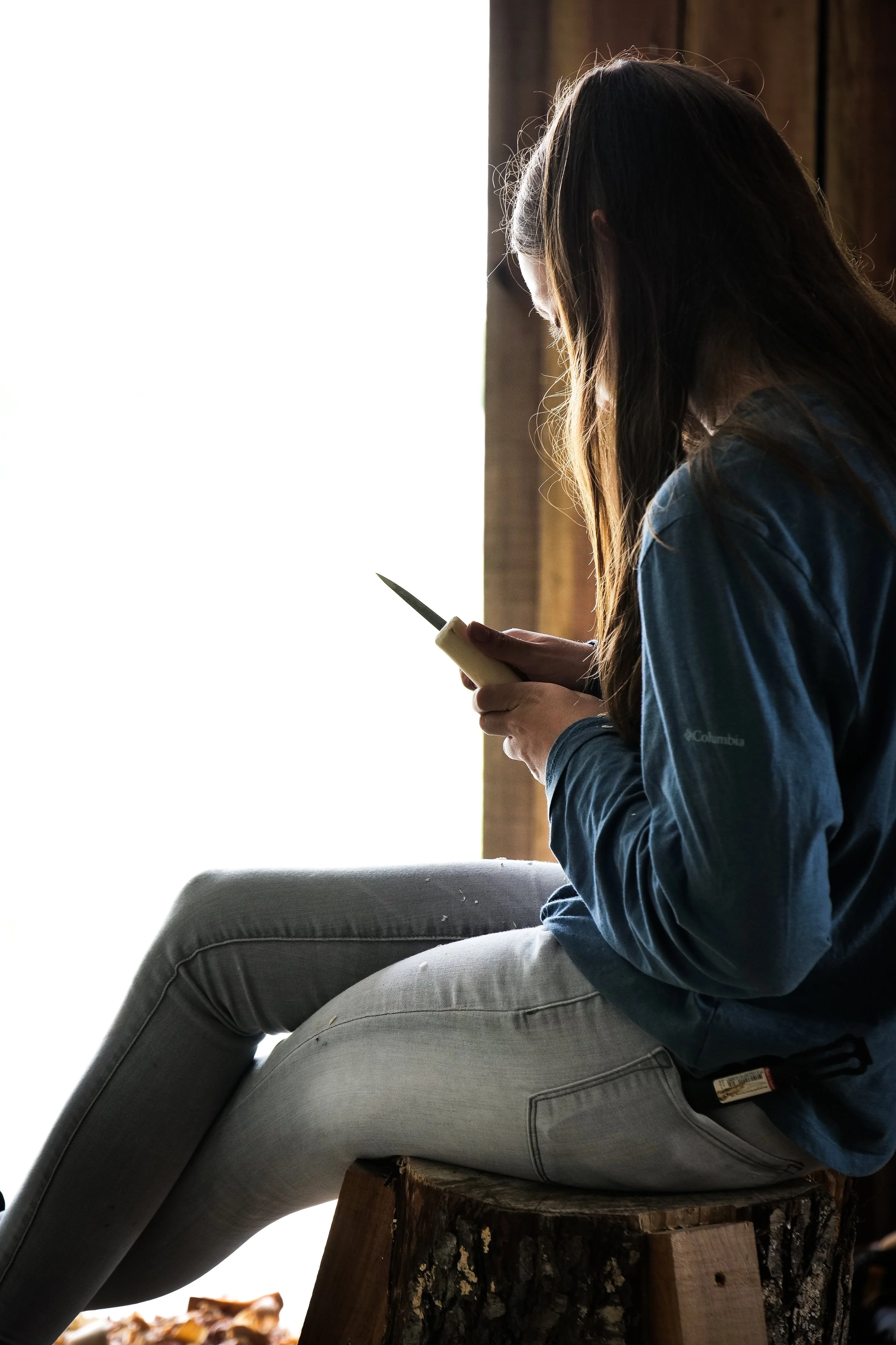 photo by Ben Aguilar. A female in jeans and long-sleeved blue t-shirt sits in profile, face to the left. She holds a knife against a piece of wood she is carving.