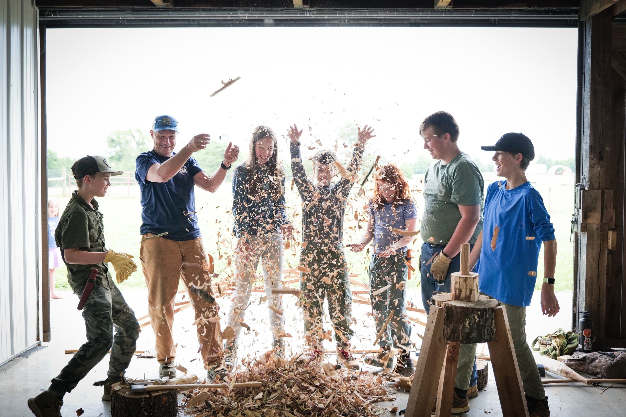 photo by Ben Aguilar. 6 middle grade kids and 1 adult toss wood shavings into the air in celebration. They are just inside the open doors of a barn; the sky is bright behind them. 