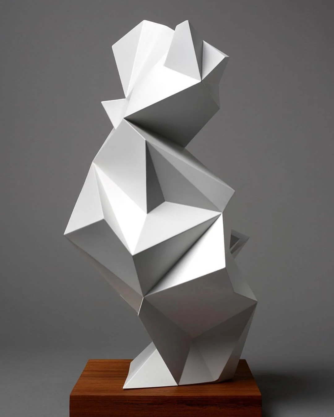 Abstract geometric sculpture with angular white and gray shapes on a wooden base, set against a gray background.