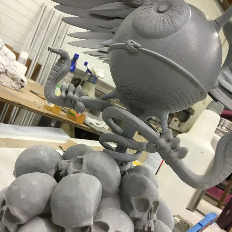 Sculpture of a surreal creature with a large orb head, one eye, snake-like arms, and a serpent-shaped tail standing on a pile of skulls in a workshop.