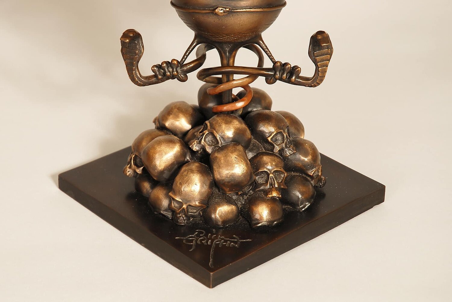 Bronze sculpture of a cross with a snake wrapped around it, positioned above a pile of skulls on a black base, signed by artist.