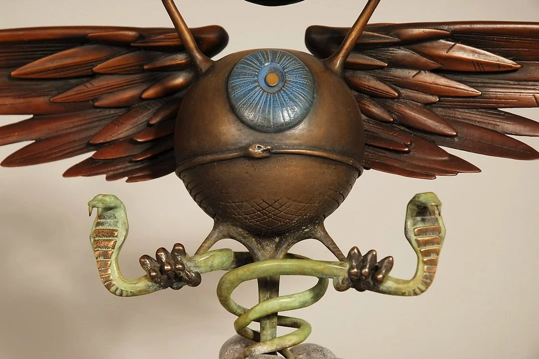 A metal sculpture of a creature with an insect-like body, large blue eye, and brown wings that resemble those of a bird or butterfly, with snake-shaped arms.