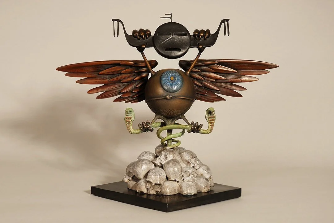 Artistic sculpture of a robot with a large blue eye, insect wings, and antennas, sitting on a pile of skulls with a jungle motif, displayed on a black base.