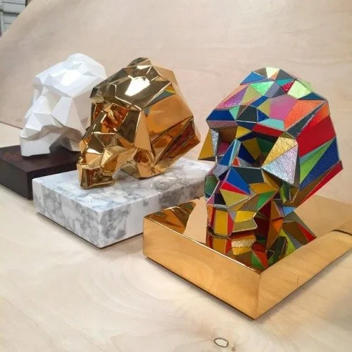 Three animal head sculptures in geometric style, each on a separate base: a white lion on a black base, a gold lion on a marble base, and a multicolored lion on a gold base.