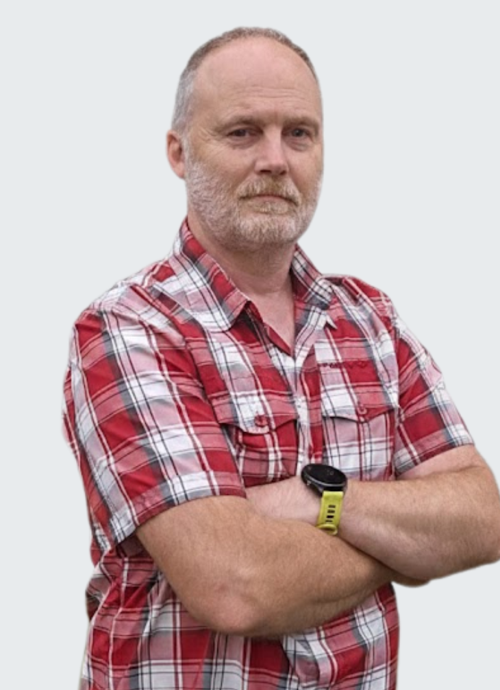 A middle-aged man with a beard and short hair, wearing a red plaid shirt and a yellow wristwatch, standing with arms crossed against a plain background.