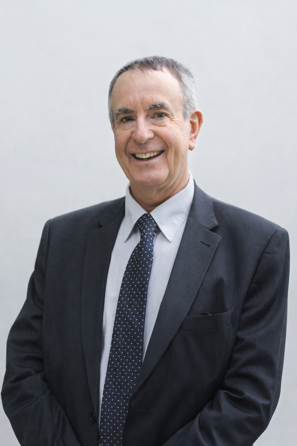 Smiling older man in a dark suit, white shirt, and dotted tie posing against a plain light background.