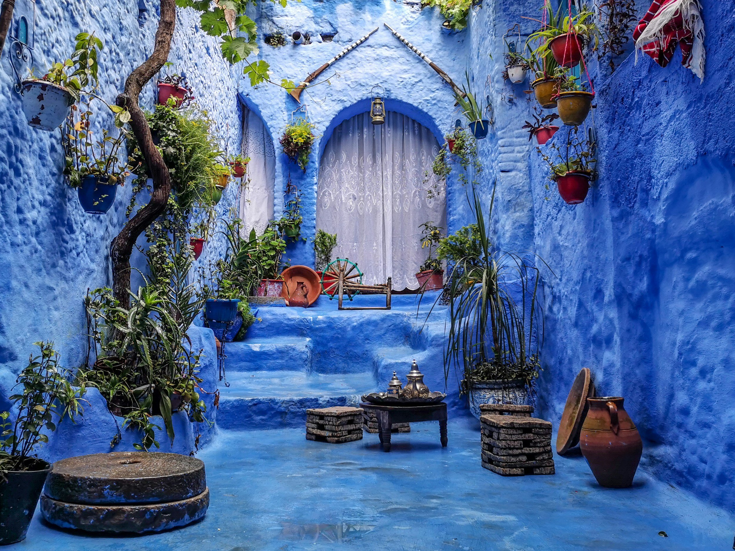 A vibrant blue courtyard with hanging flower pots, potted plants, rustic bricks, and decorative items.