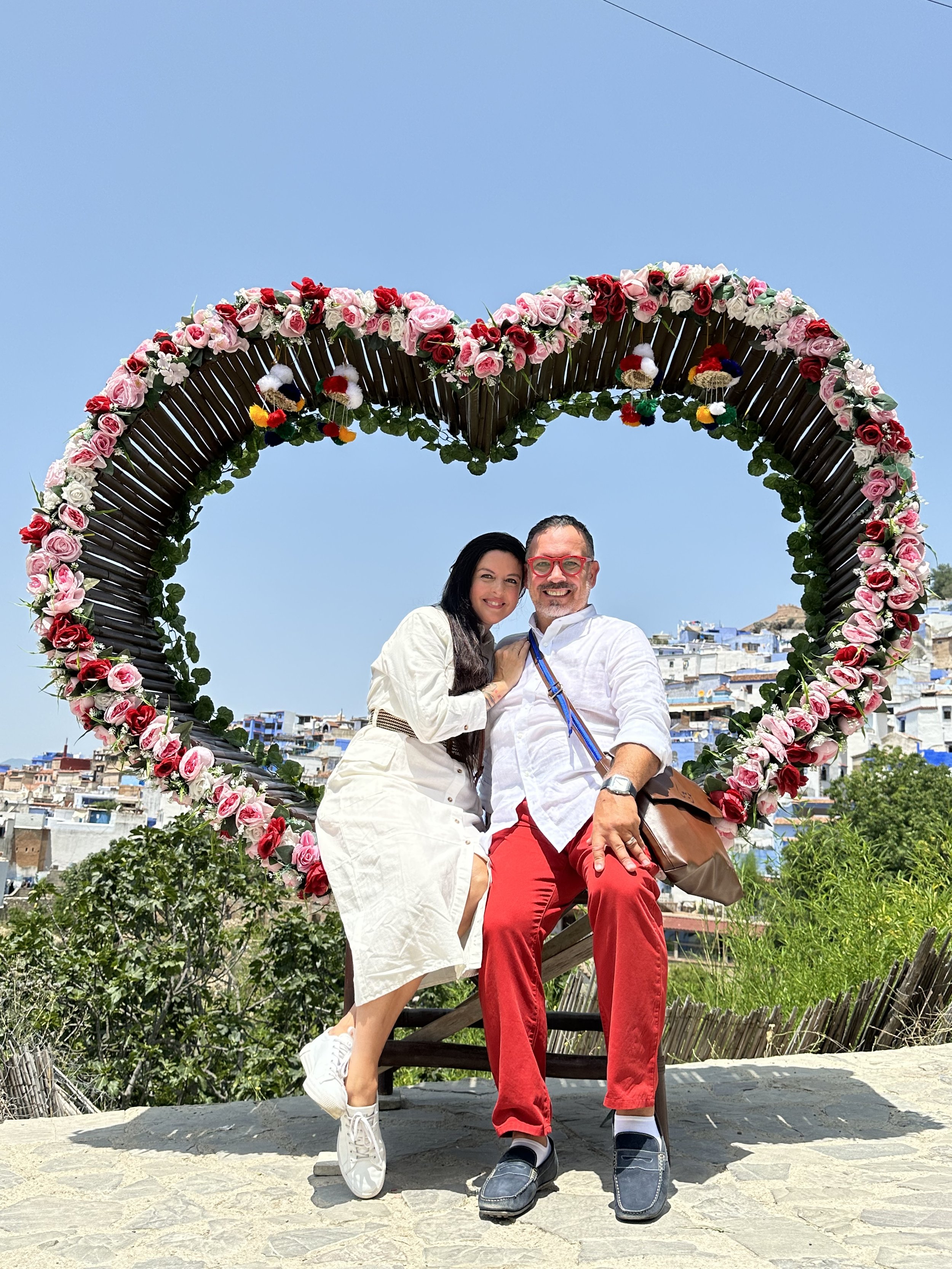 A couple sitting on a bench in front of a large heart-shaped floral decoration with pink and red flowers. They are smiling, dressed casually, with a cityscape in the background under a clear blue sky.
