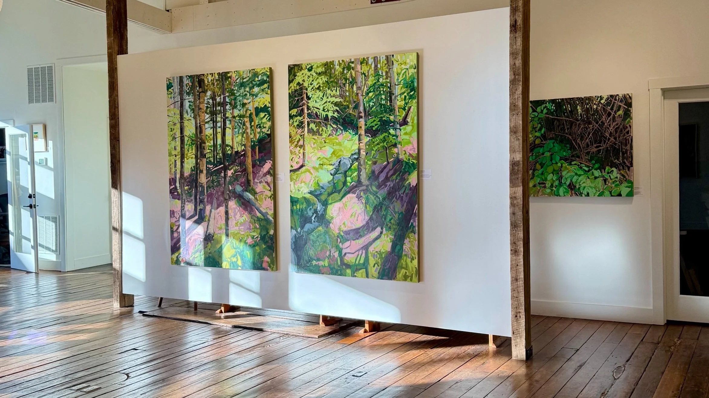 A gallery photo of two large tree paintings centered on the wall. Behind the wall is another painting. The sun shines in on the walls.