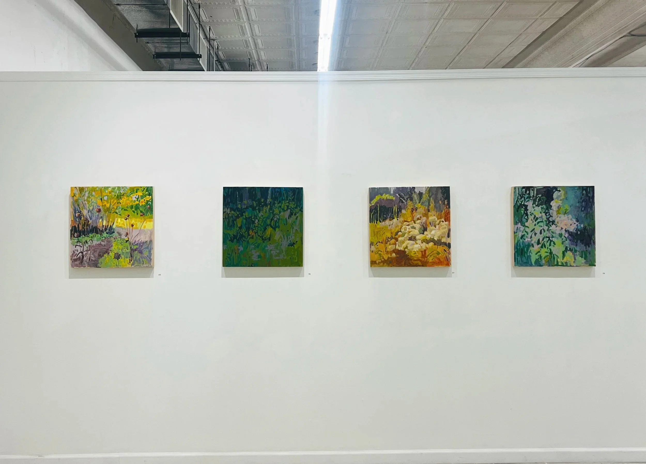 Four square paintings evenly spaced hanging on a white wall. The paintings are all abstract landscapes.