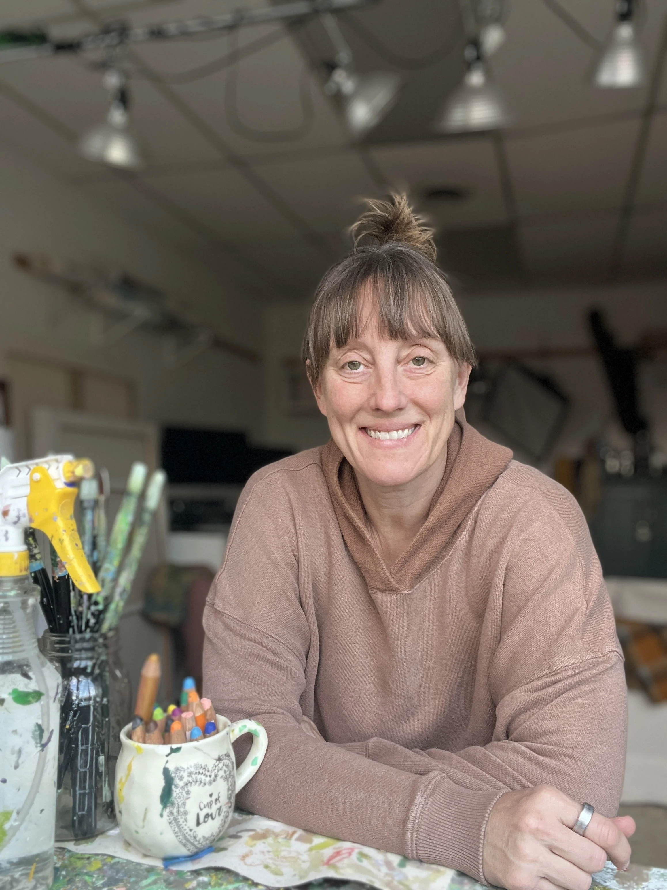 A photo of Krista smiling at the camera while leaning on her work table in her studio. The studio and art supplies can be seen around her.