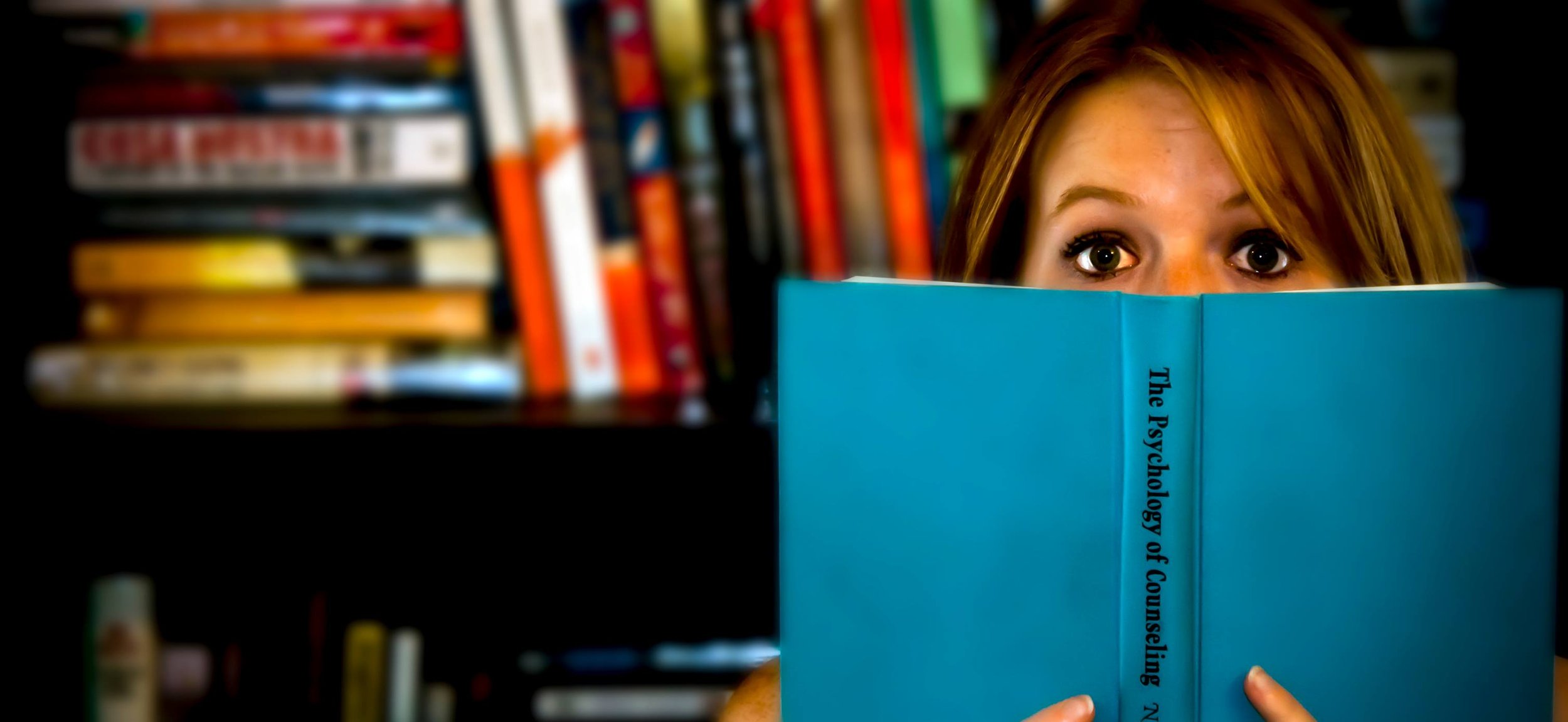 A woman with reddish-brown hair and wide eyes is hiding behind a blue book titled 'The Psychology of Counseling.' She is in a library, with shelves of books in the background.