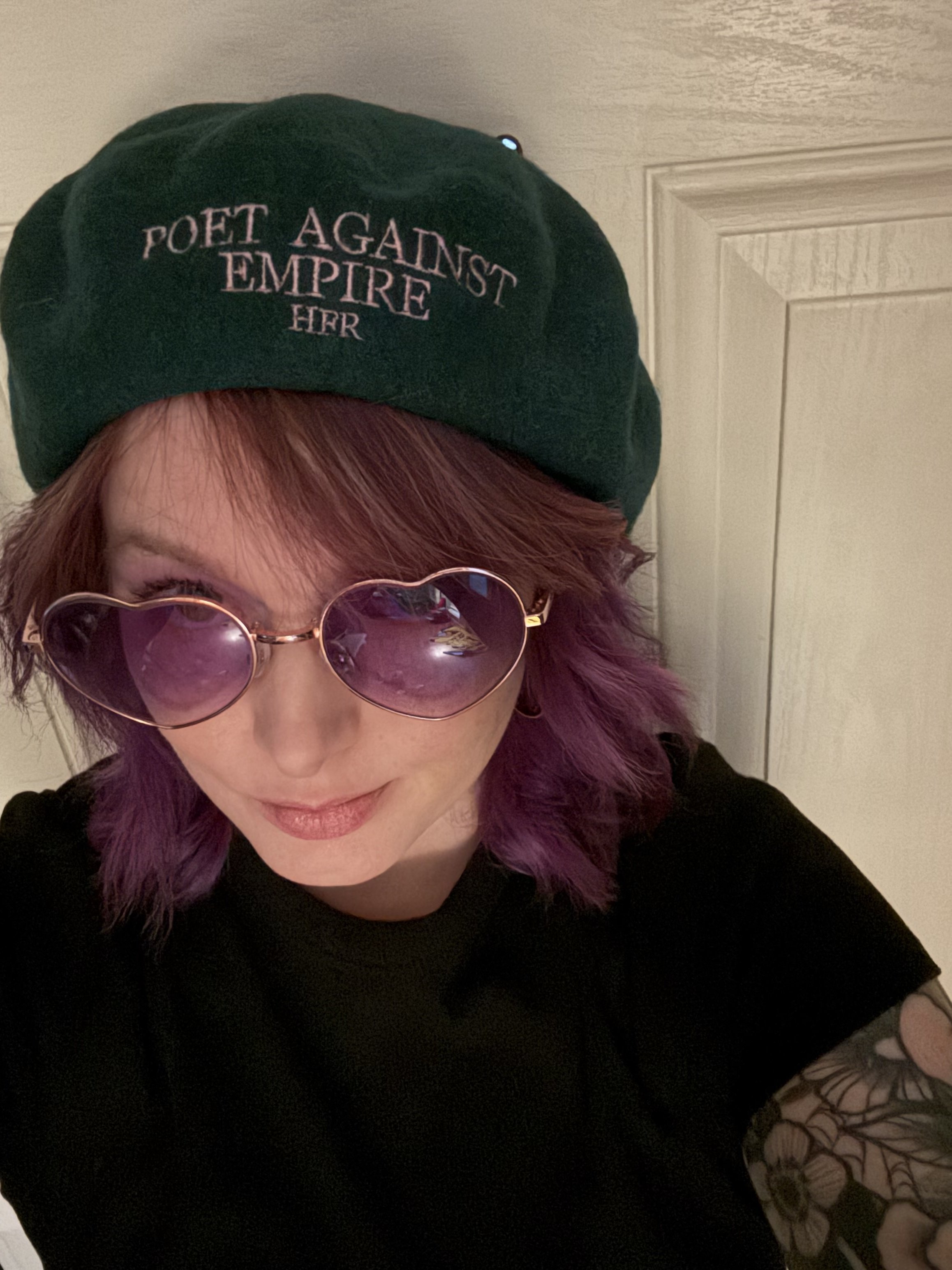 A woman with short, reddish hair wearing heart-shaped sunglasses, a black shirt, and a black beret embroidered with the phrase 'Poet Against Empire' in pink. She is standing in front of a beige door.