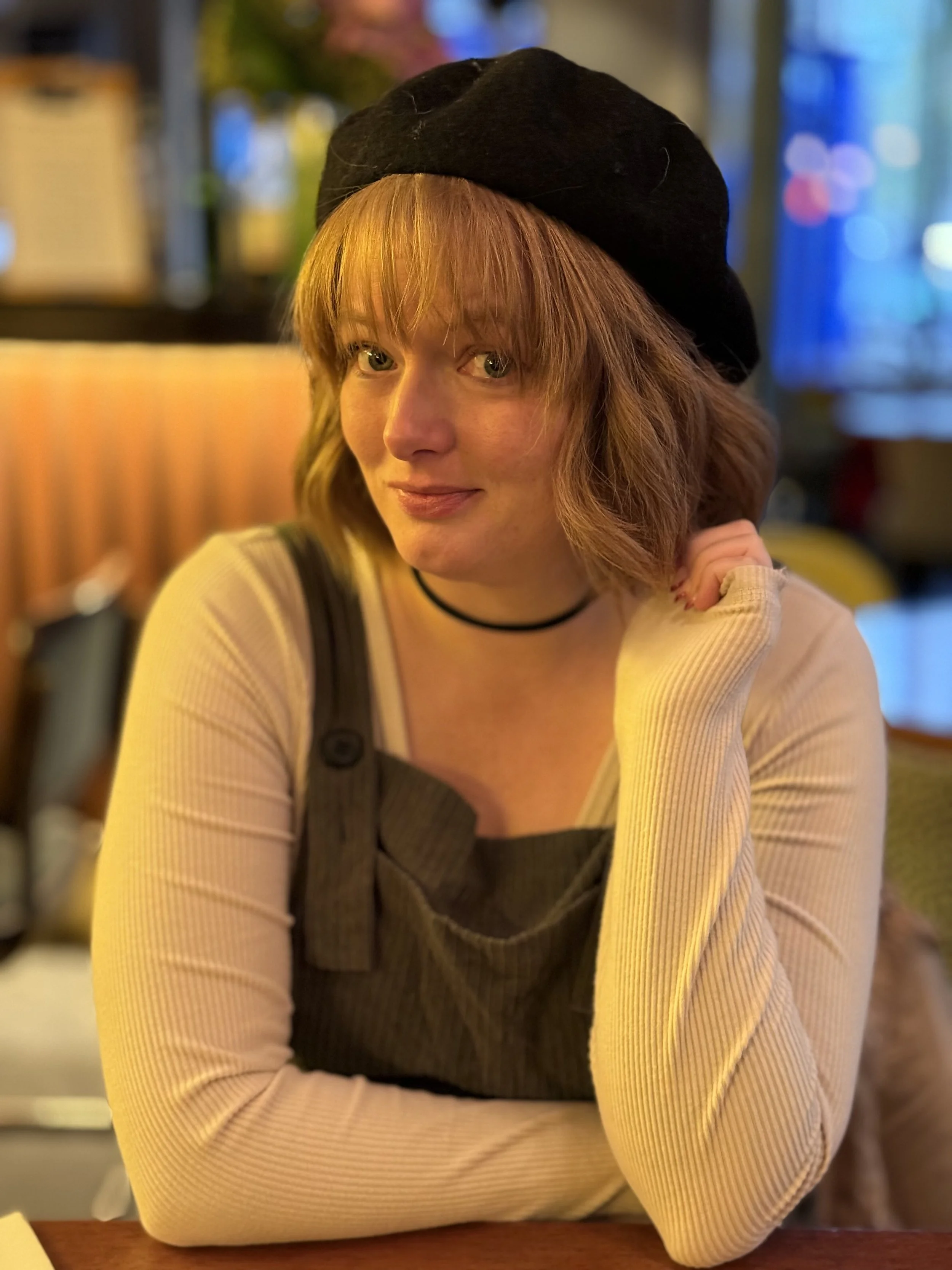 A woman with shoulder-length auburn hair and green eyes, wearing a beige long-sleeve top and a black beret, sitting in a warmly lit indoor space with a blurred background of colorful lights.