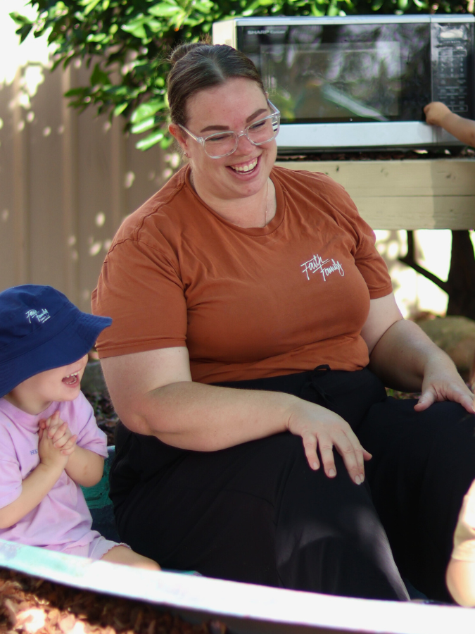 A woman with glasses smiling and sitting outdoors next to a young girl wearing a blue hat. They are enjoying a moment together, with a microwave visible in the background.