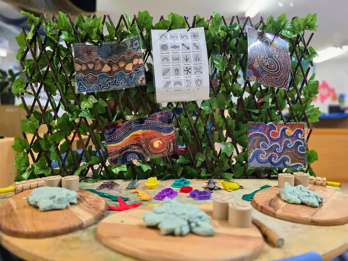 Table with playdough on wooden boards, rollers, stamps and cutters with a display of indigenous artworks and symbols