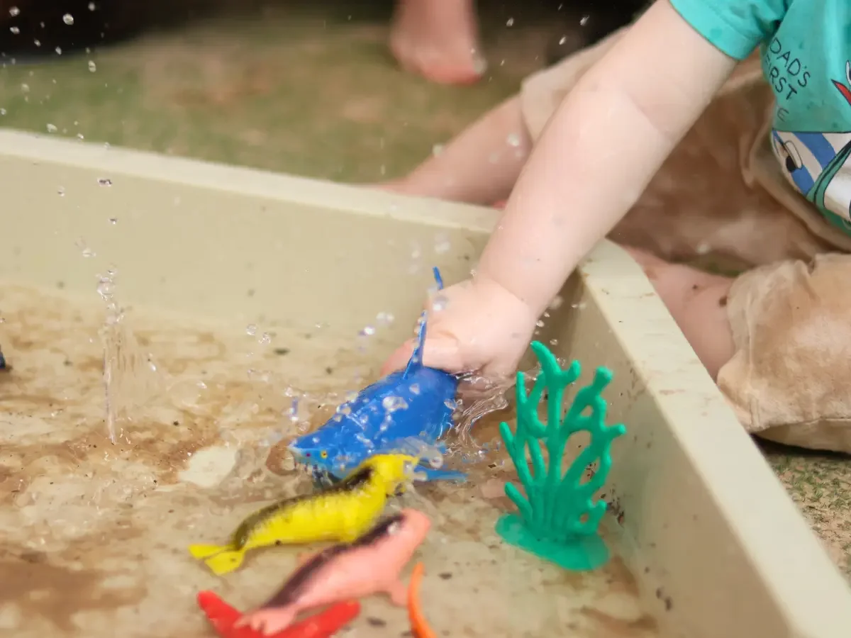 Child holding a figurine of a shark splashing it in a tray of water with other ocean animals