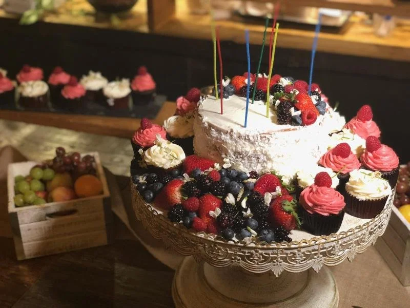 A decorated birthday cake with a white frosted cake with fruit toppings and colorful candles, surrounded by pink and white cupcakes with raspberries, on a cake stand.