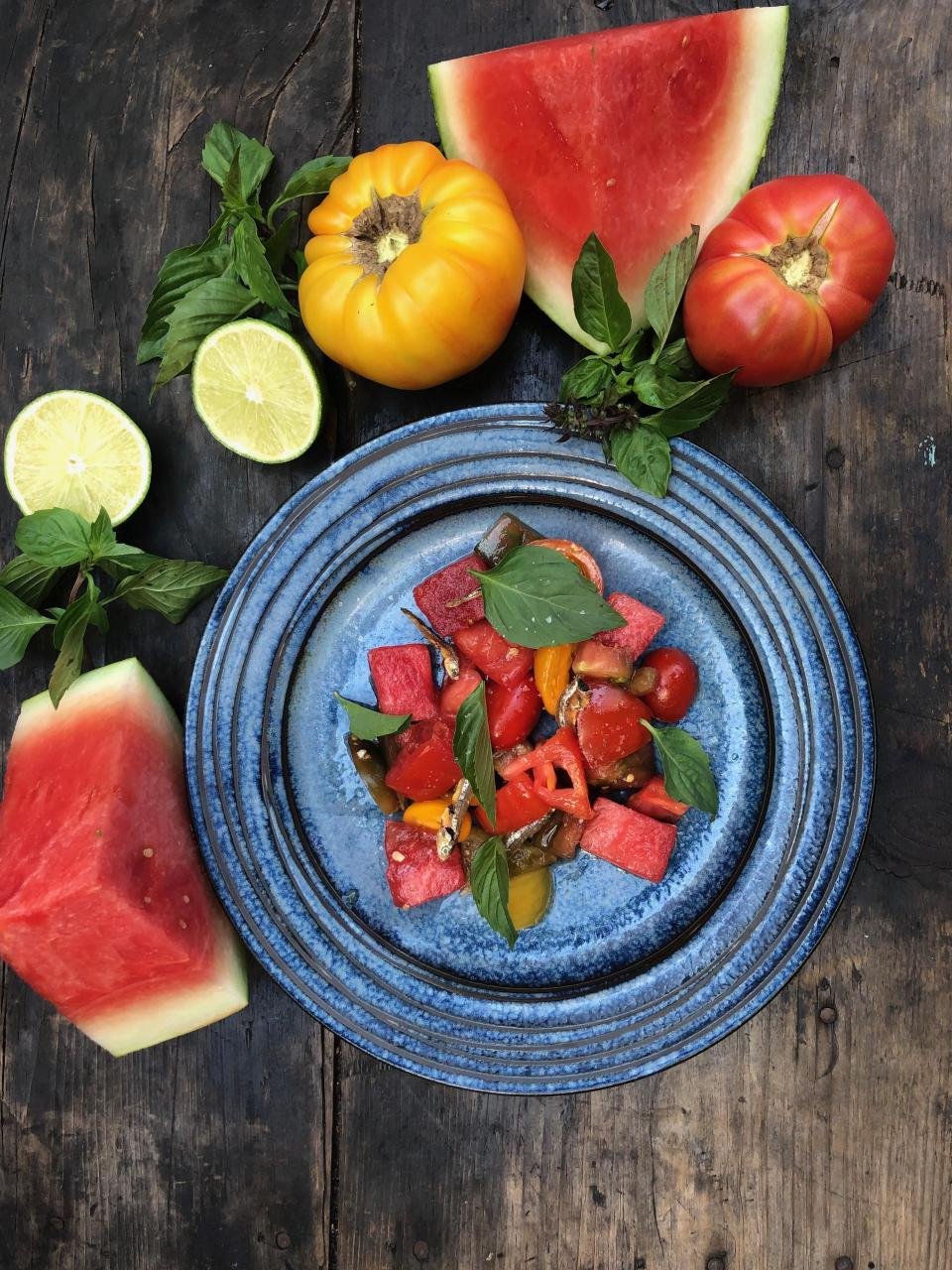 Plate of chopped watermelon, cherry tomatoes, and basil leaves on a rustic wooden table, surrounded by lemon halves, slices of watermelon, and tomatoes.