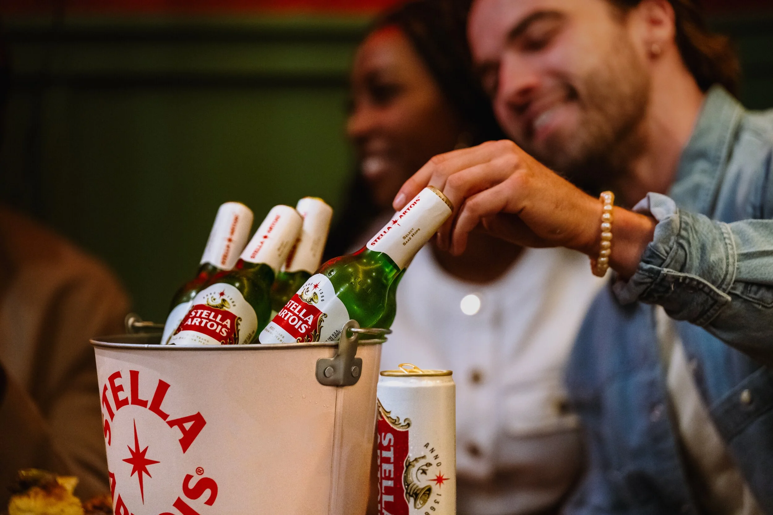 A person with long hair, wearing a denim jacket and a beaded bracelet, reaching into a Stella Artois beer bucket filled with bottles, with smiling people in the background.