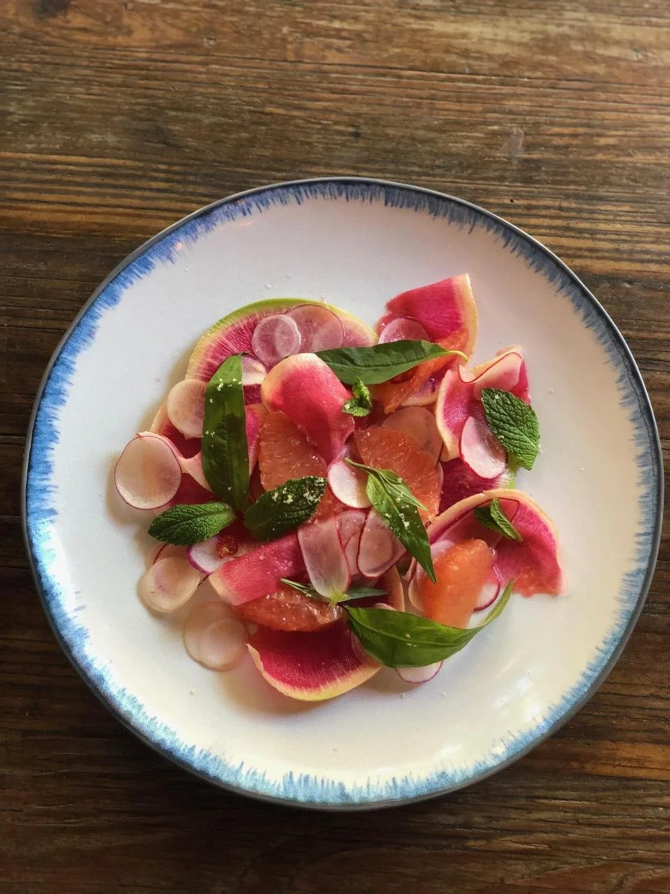 Salad with sliced watermelon radish, grapefruit, thinly sliced radish, and fresh mint and basil leaves on a white plate with blue rim, placed on a wooden surface.