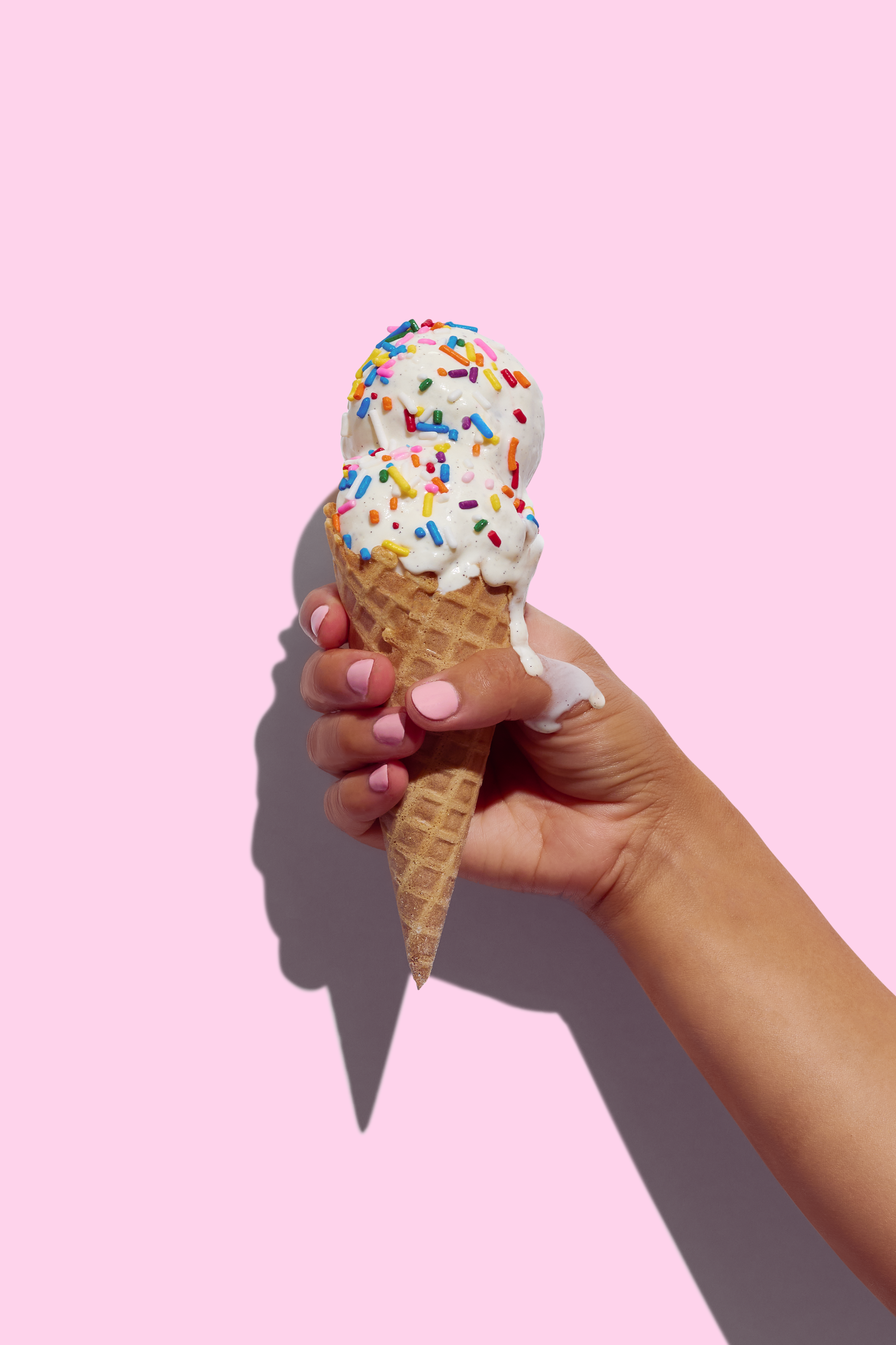 A hand holding a vanilla ice cream cone with colorful sprinkles on a pink background.