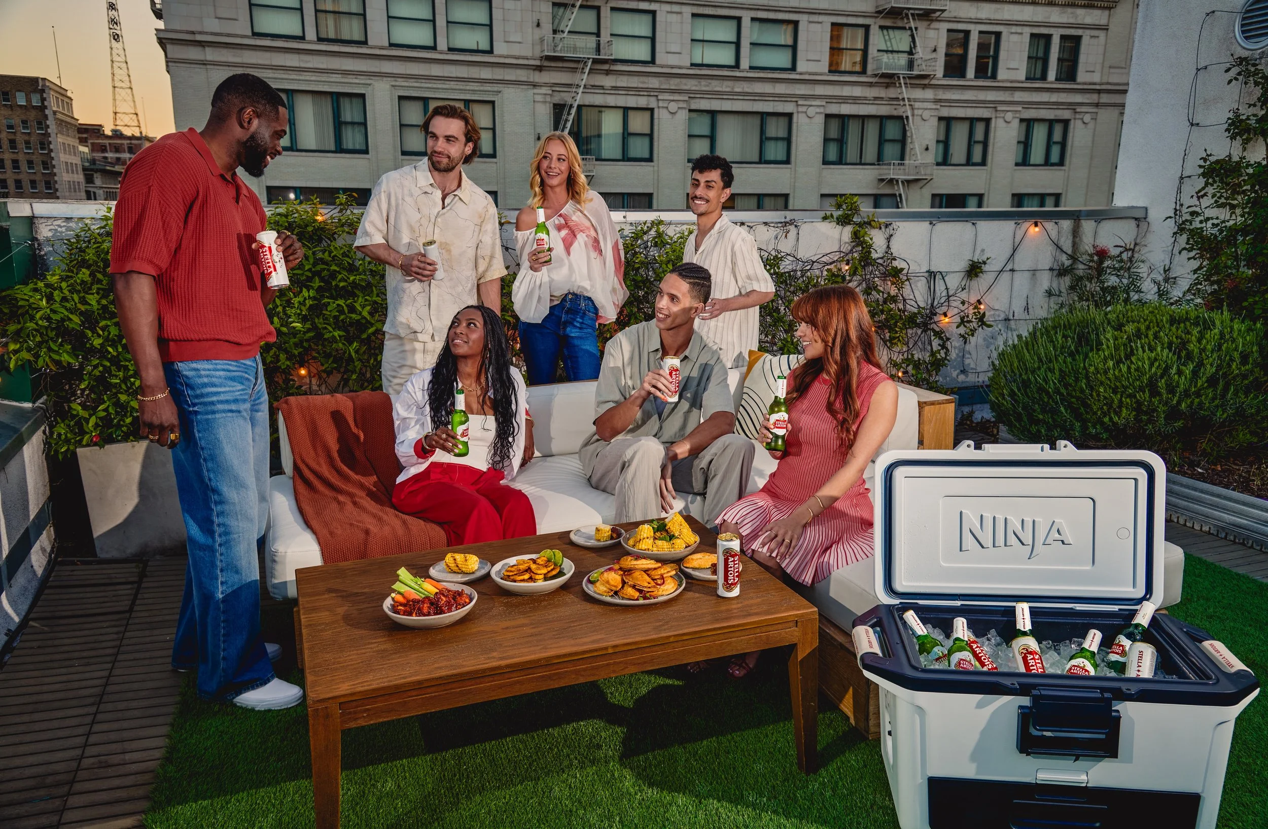 Group of eight friends enjoying drinks and snacks on a rooftop terrace with city buildings in the background, seated on a couch and standing around a table with food, with a cooler filled with beers nearby.