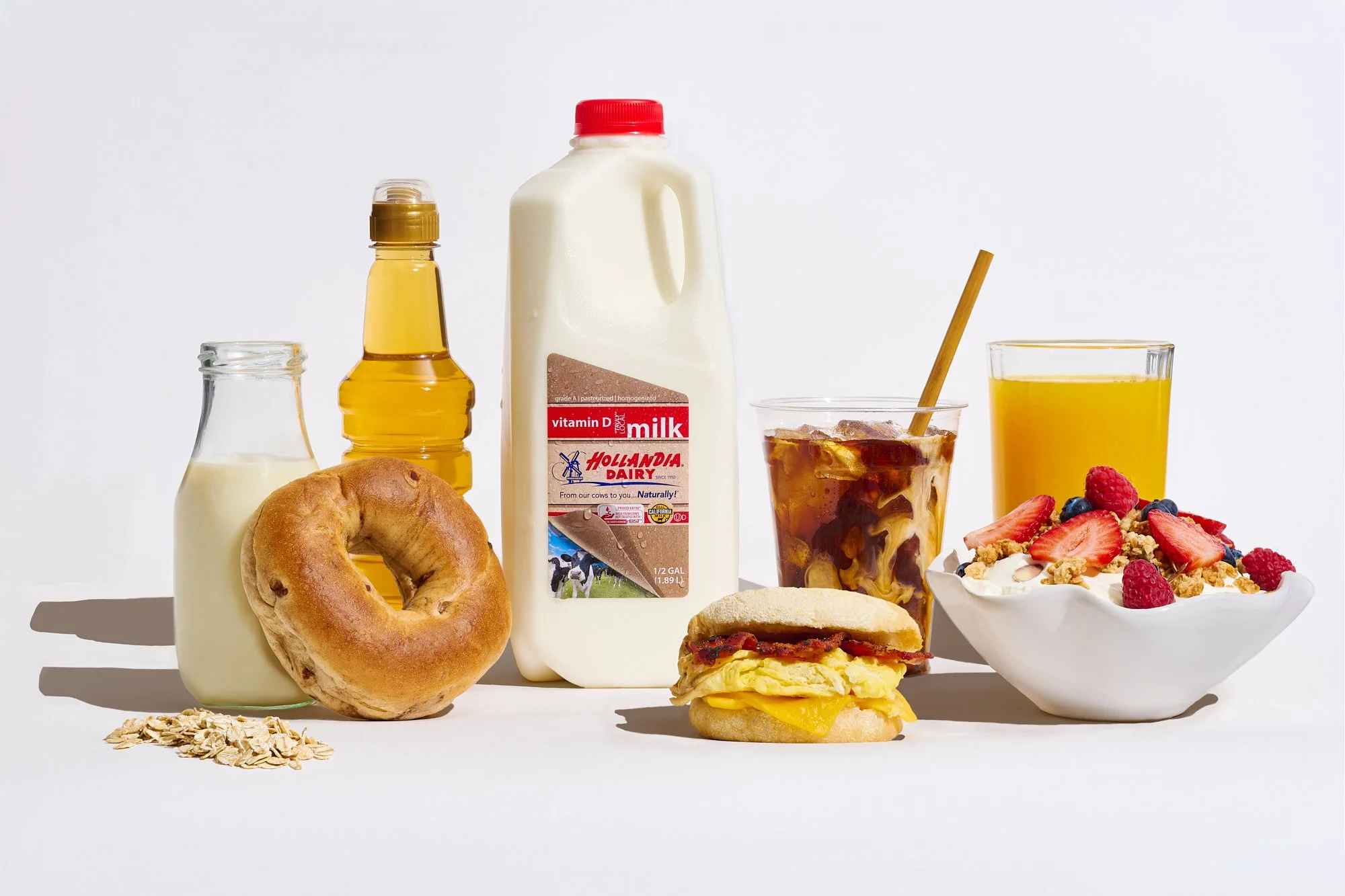 Breakfast spread with milk, orange juice, iced coffee, bagel, oats, breakfast sandwich, and a bowl of yogurt with strawberries and raspberries.