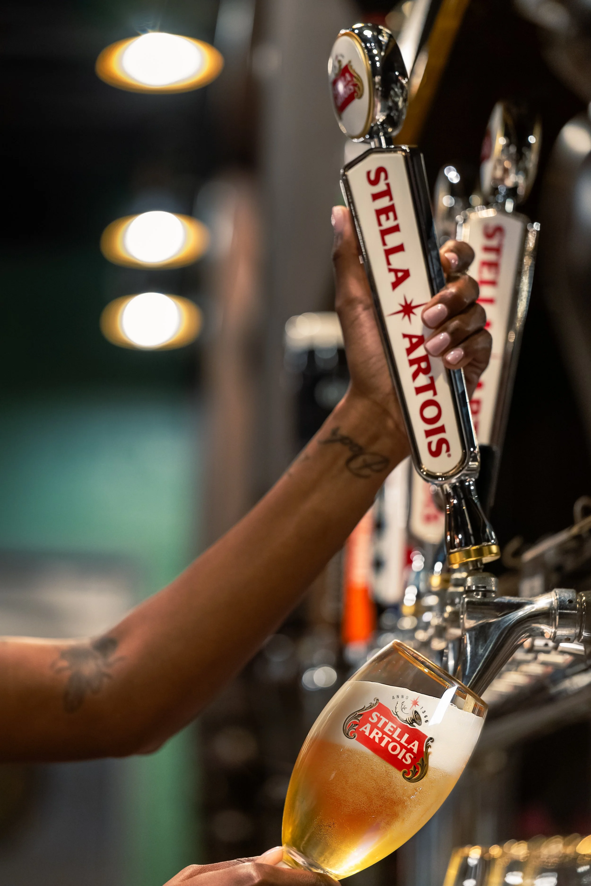 A person pouring Stella Artois beer from a tap into a glass with Stella Artois logo. The person's hand is visible, with a tattoo on the wrist. The background is blurred with circular lights.