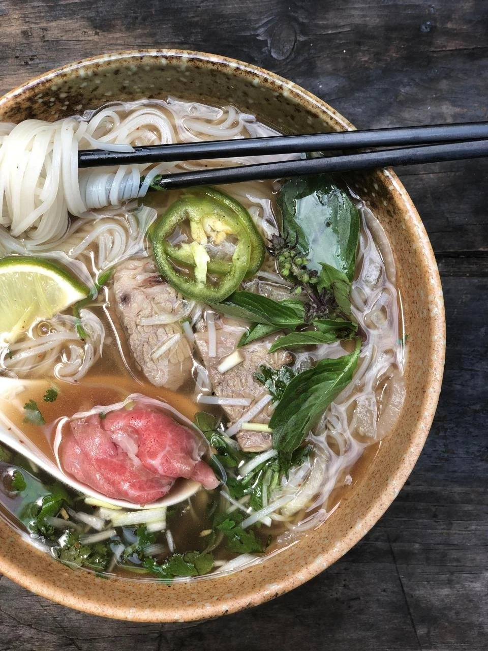 Bowl of Vietnamese Pho with rice noodles, beef slices, basil, lime, jalapeño, and fresh herbs on rustic wooden table, chopsticks resting on the bowl.