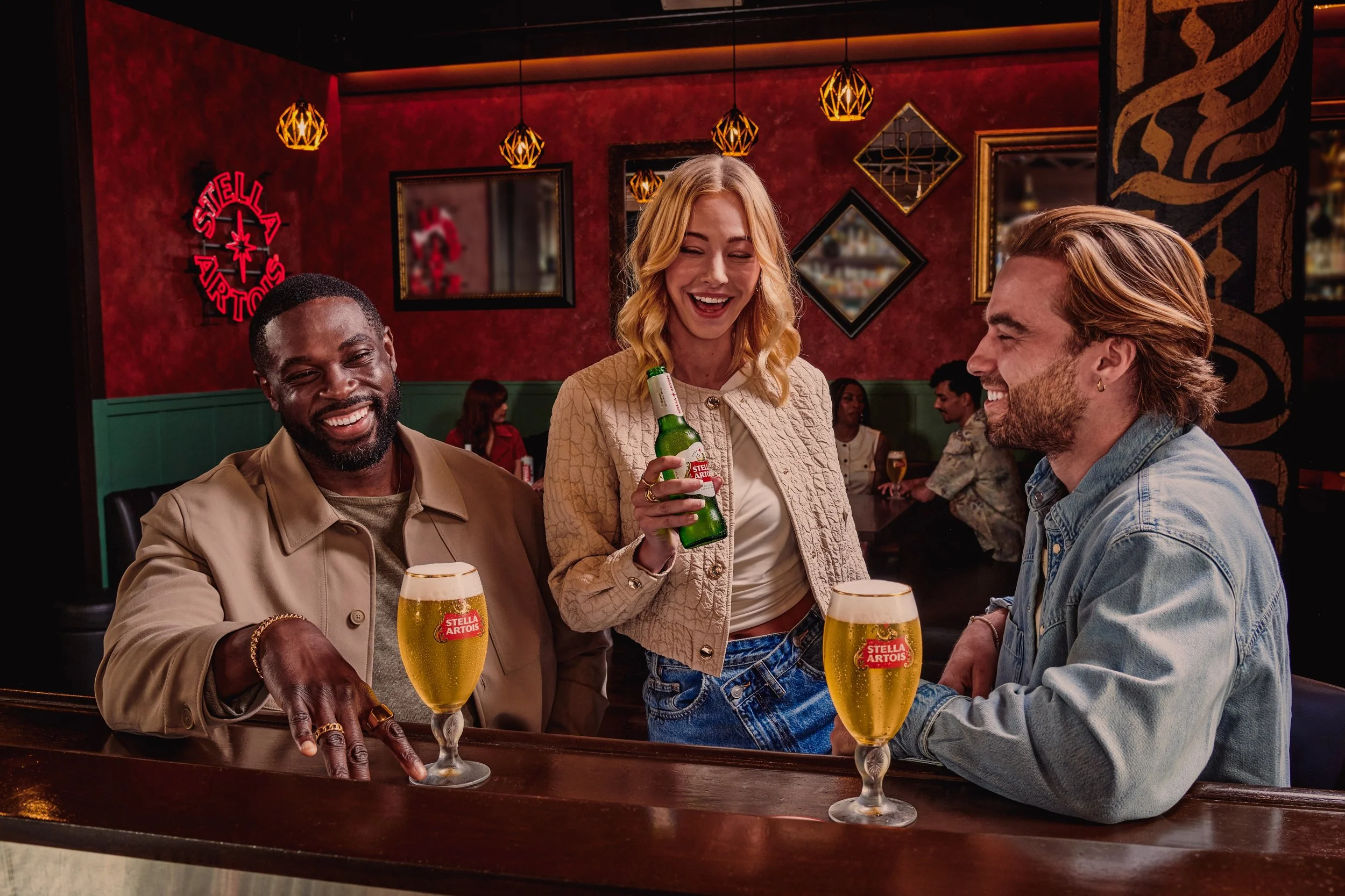 Three friends enjoying drinks at a bar, with two glasses of Stella Artois beer on the counter and a woman holding a beer bottle, all smiling and laughing.