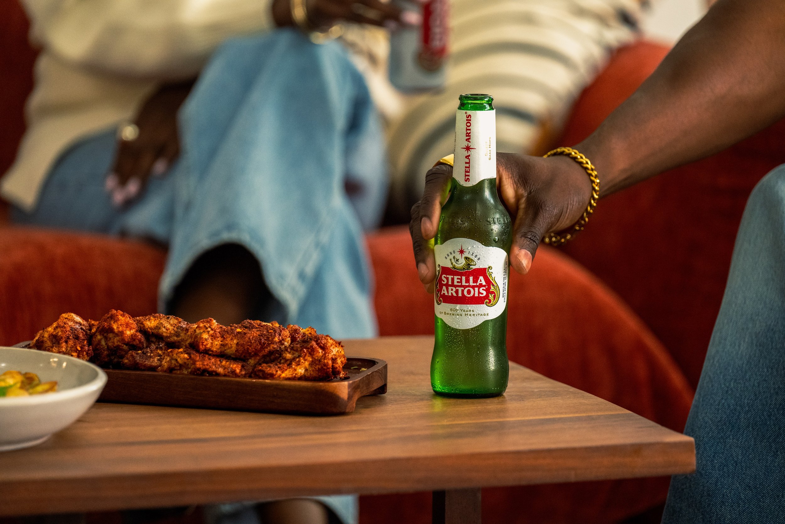 Hand holding a Stella Artois beer bottle on a wooden table, with a plate of grilled meat and a bowl of food nearby. In the background, a person is sitting on an orange couch wearing a yellow and white striped shirt and blue jeans.