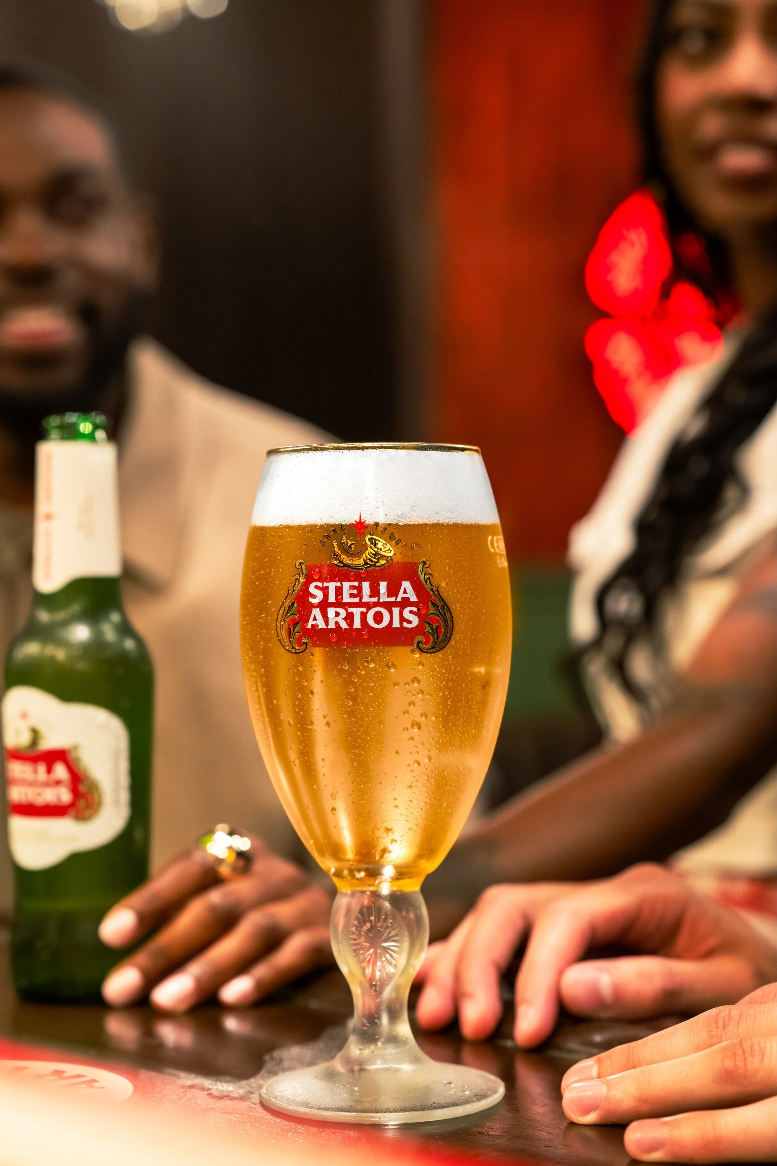 A glass of Stella Artois beer with condensation on it, placed on a bar counter. Two people's hands are visible nearby, one with a ring, and a green beer bottle in the background. The scene appears to be in a bar or pub setting, with two individuals p