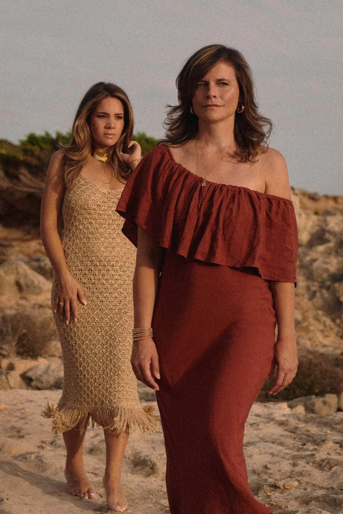 Two women walking on a beach during sunset, one in a off-shoulder red dress and the other in a beige dress with fringes at the bottom.