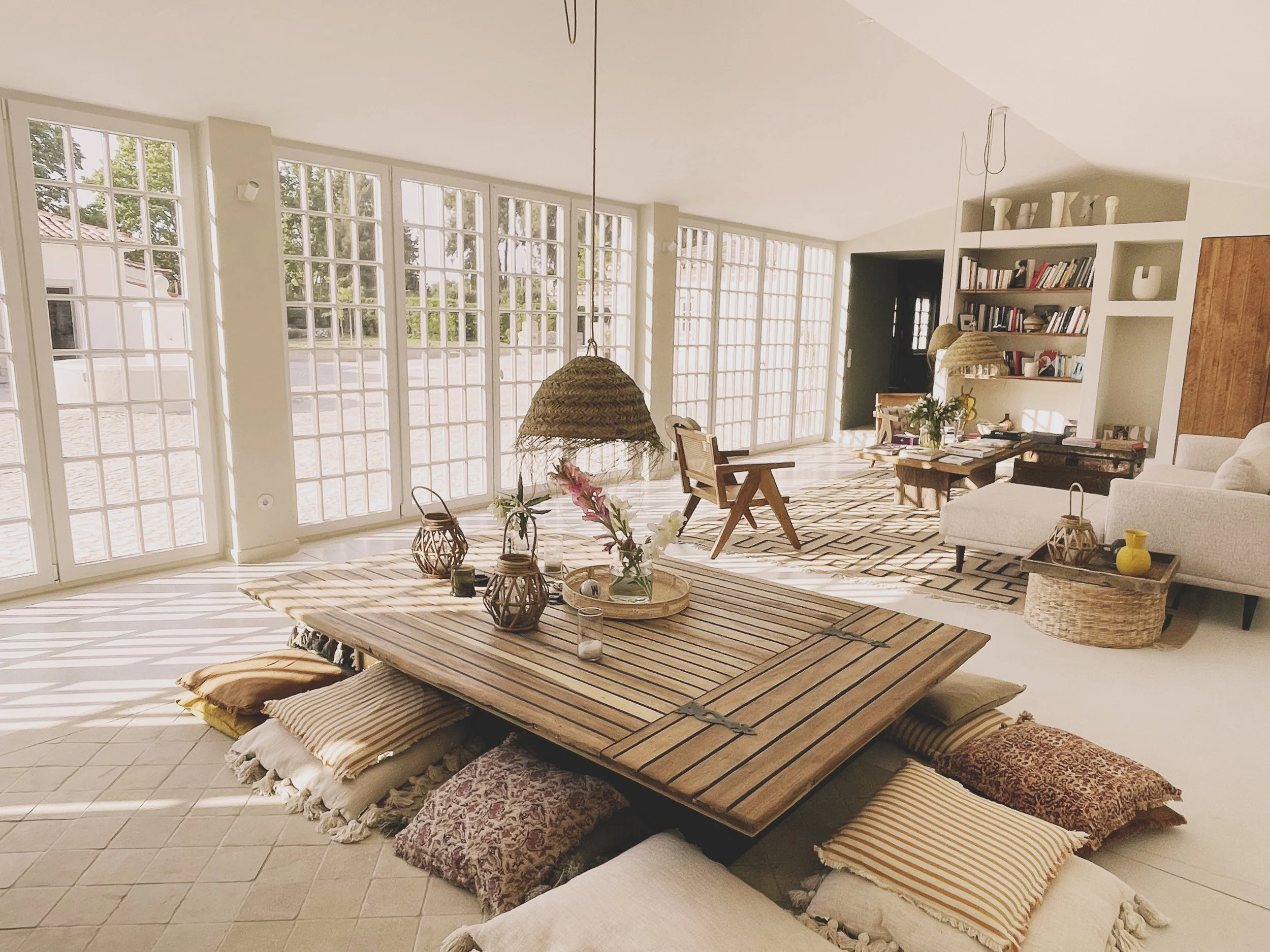Bright living room with large grid-style windows, a wooden low table surrounded by floor cushions, and a bookshelf with decorative vases and books.
