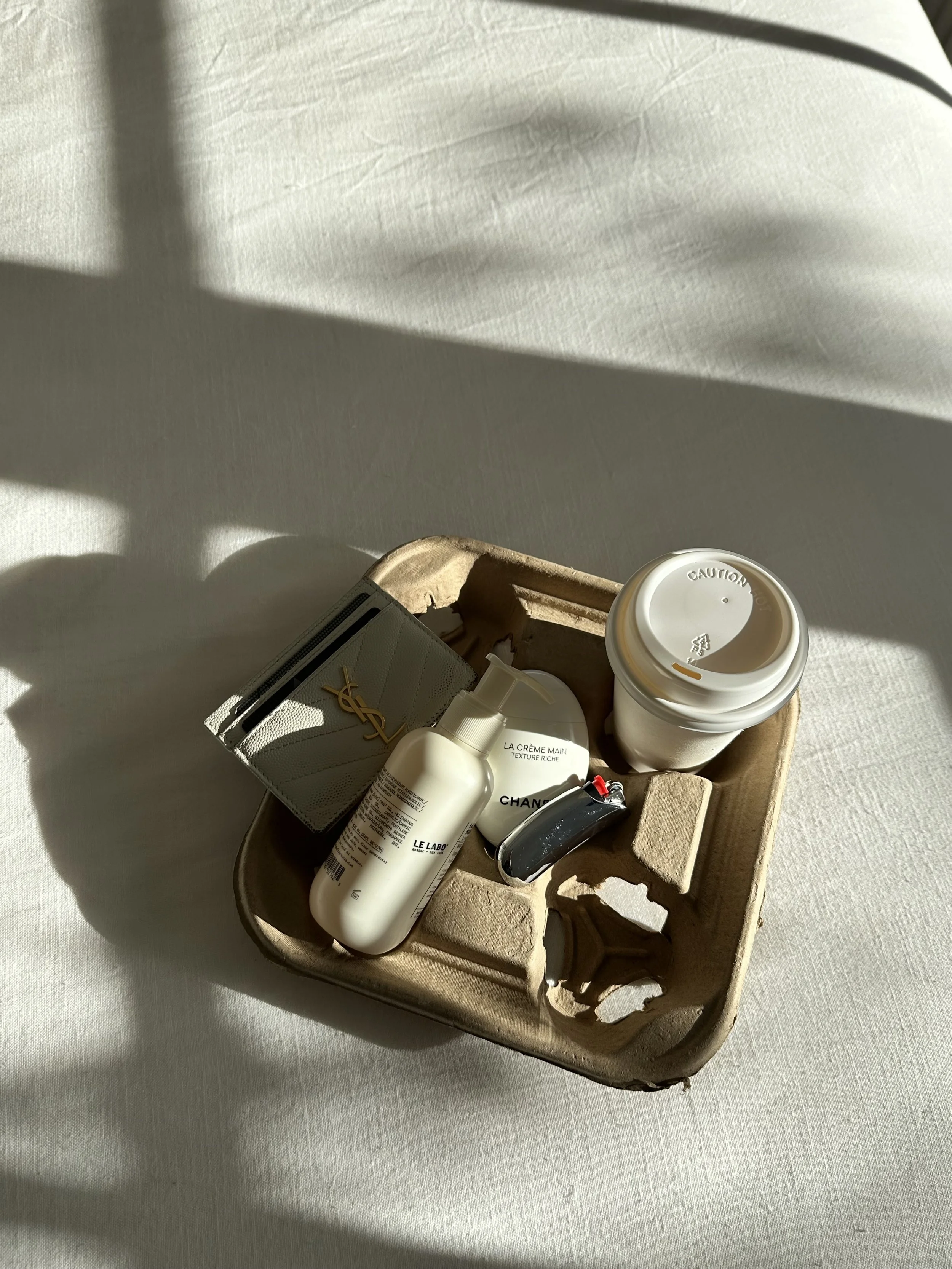 A cardboard tray holding several personal care and beauty products, including a white YSL cosmetic bag, a Chanel container, a La Crème Main hand cream, a Le Labo bottle, a coffee cup with a lid, and other small items, on a white surface with sunlight creating shadows.