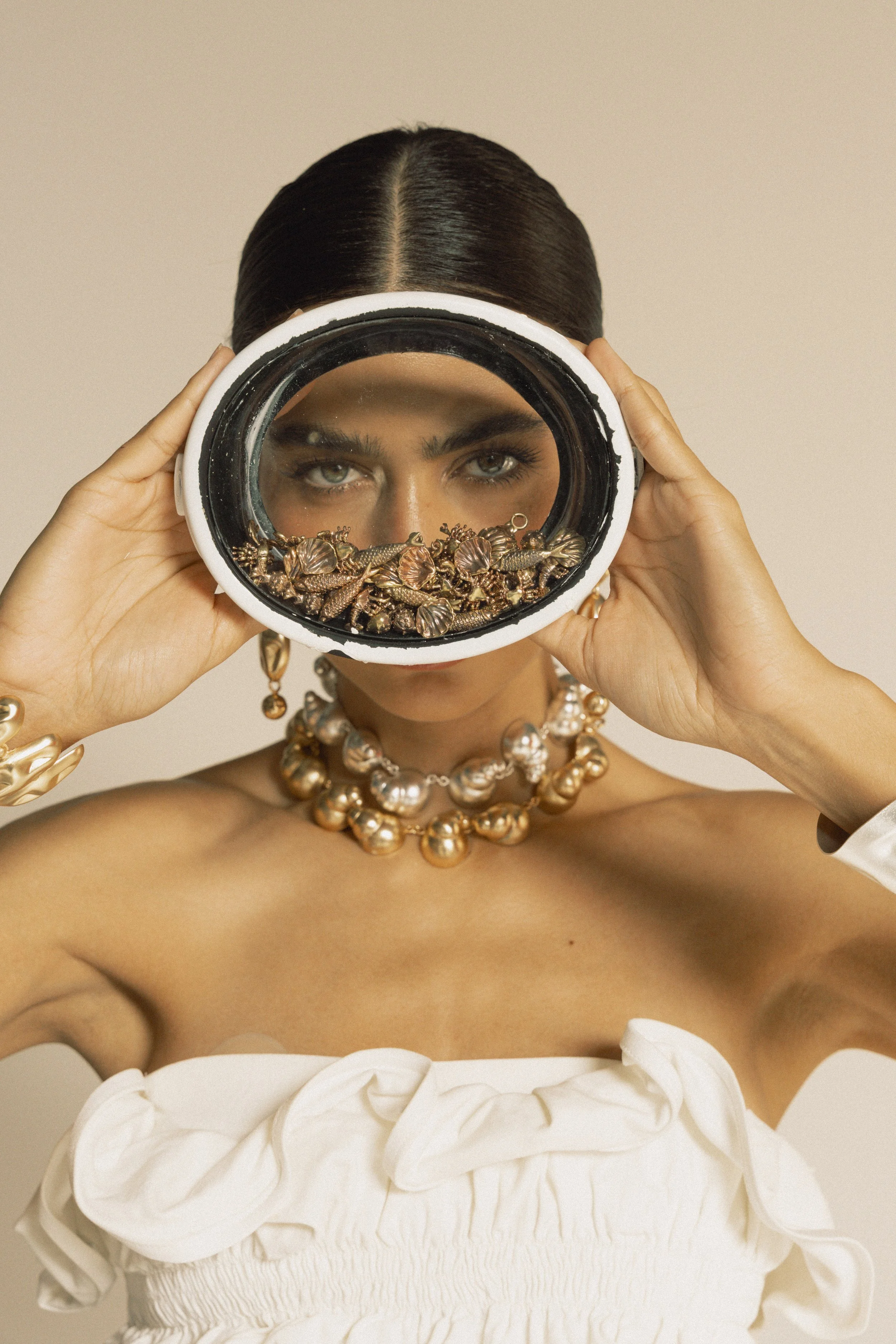 A woman with dark hair and gray eyes looking through a circular mirror she is holding up to her face, reflecting her eyes and eyebrows. She is wearing gold jewelry including earrings, a necklace, and a bracelet, and has a white, ruffled strapless dress.