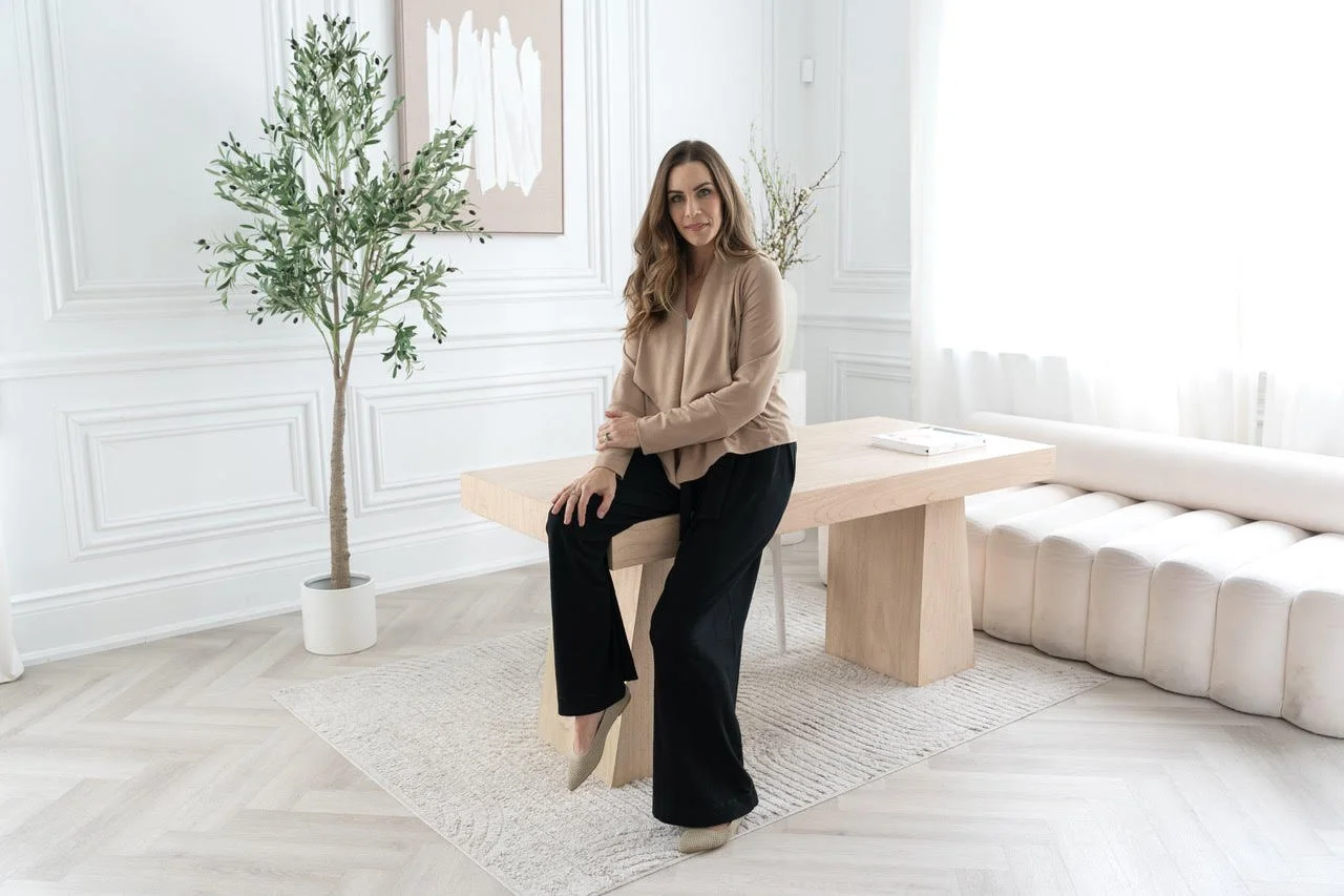 A woman sitting on the edge of a wooden table in a bright, modern living room with white walls, large windows with sheer curtains, a potted plant, and a white sofa.