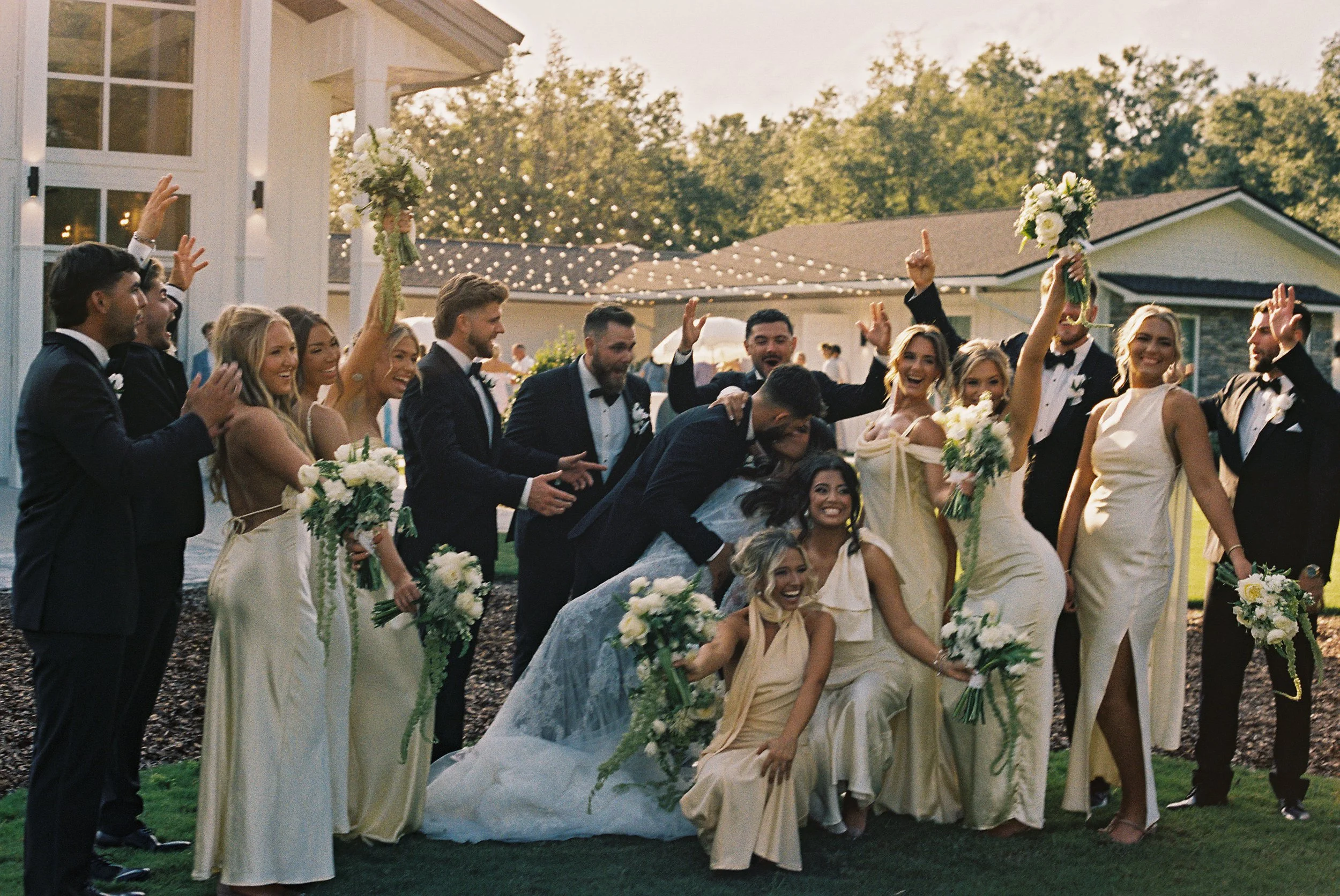 A large group of wedding guests, including men in tuxedos and women in light-colored dresses, are gathered outdoors. Some are holding bouquets and are smiling and celebrating, with a house and trees visible in the background, under string lights at s