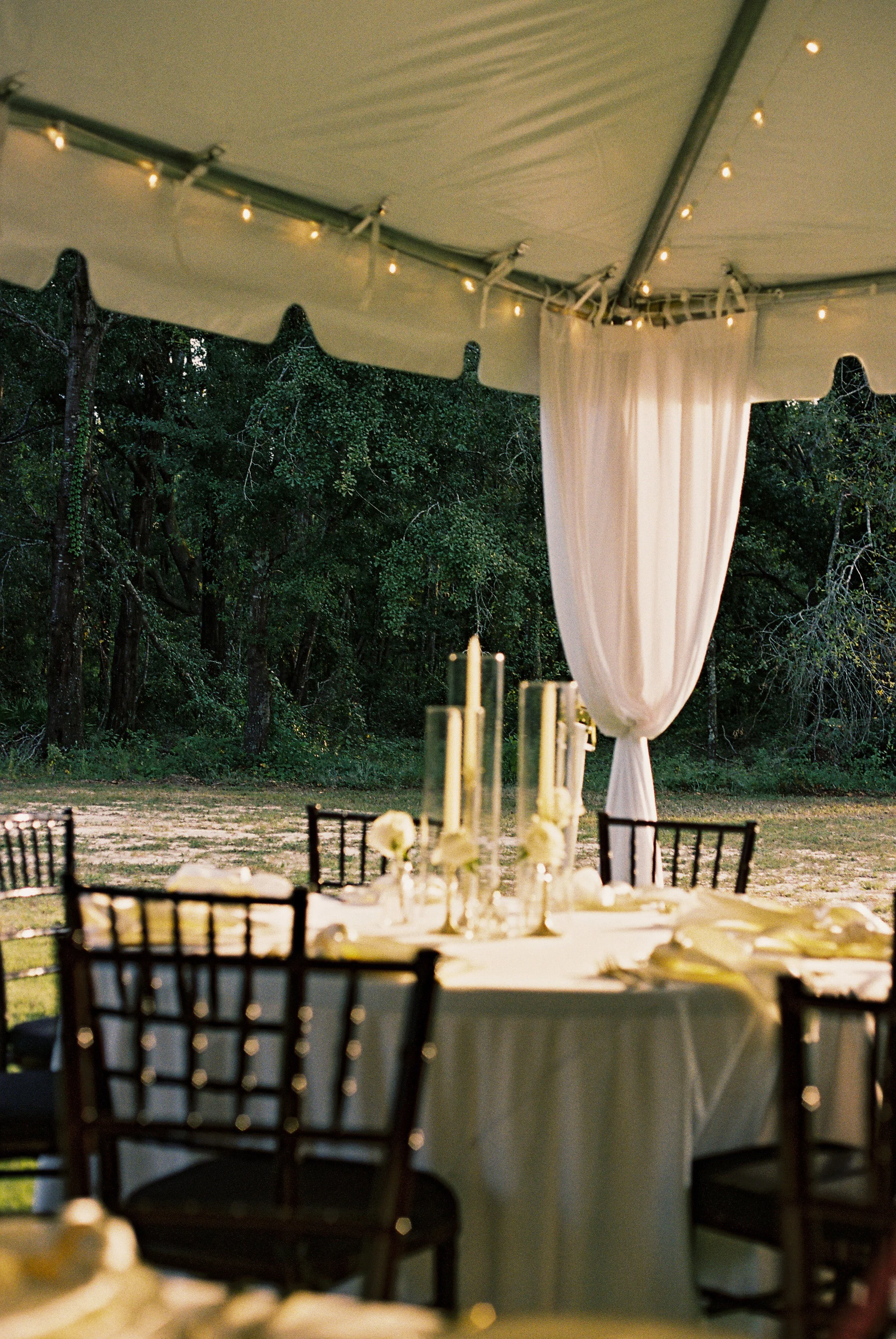 Outdoor wedding reception setup beneath a tent with string lights, round tables with white tablecloths and floral centerpieces, and black chairs, surrounded by trees and greenery.