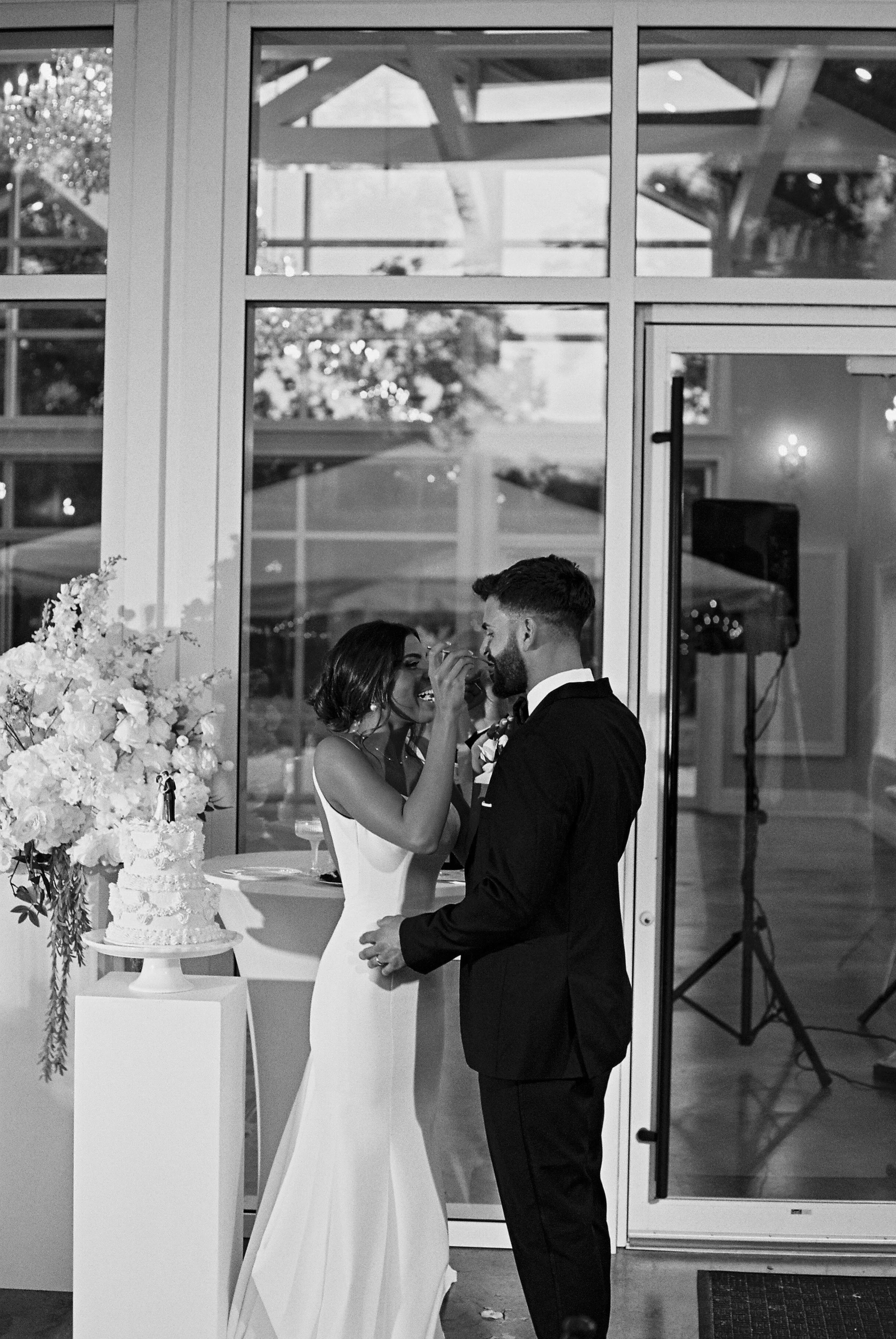 A bride and groom sharing a moment during their wedding inside a room with large glass windows, a wedding cake on a table, and floral arrangements.