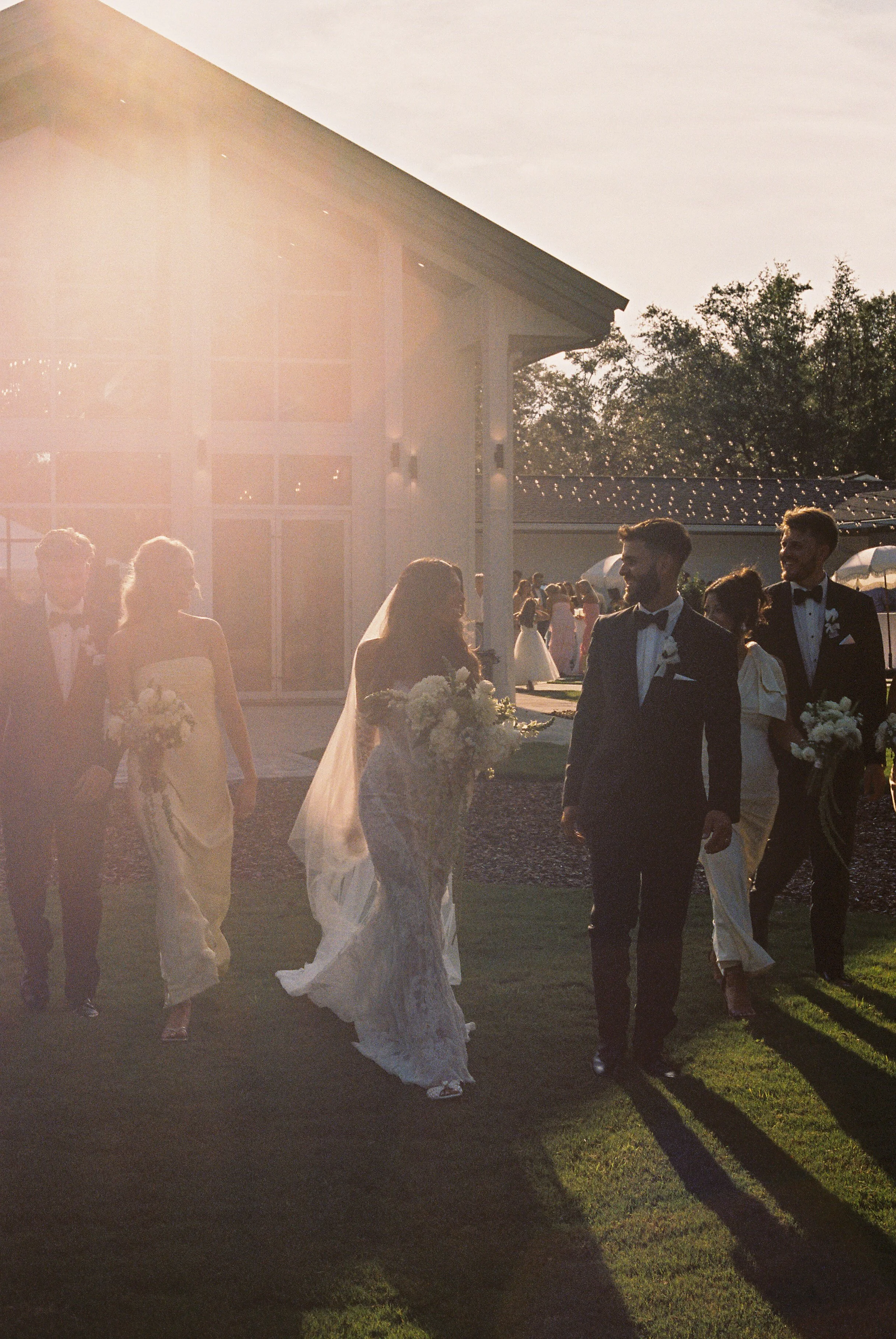 Wedding guests dressed in tuxedos and wedding dresses, walking outdoors in sunlight, with a barn-like building and string lights in the background.