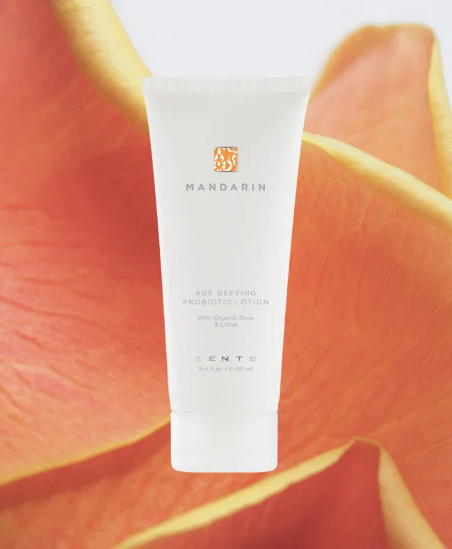 A white tube of Mandarín Age Defying Probiotic Lotion with Organic Shea and Lotus, placed in front of a background of orange flower petals.