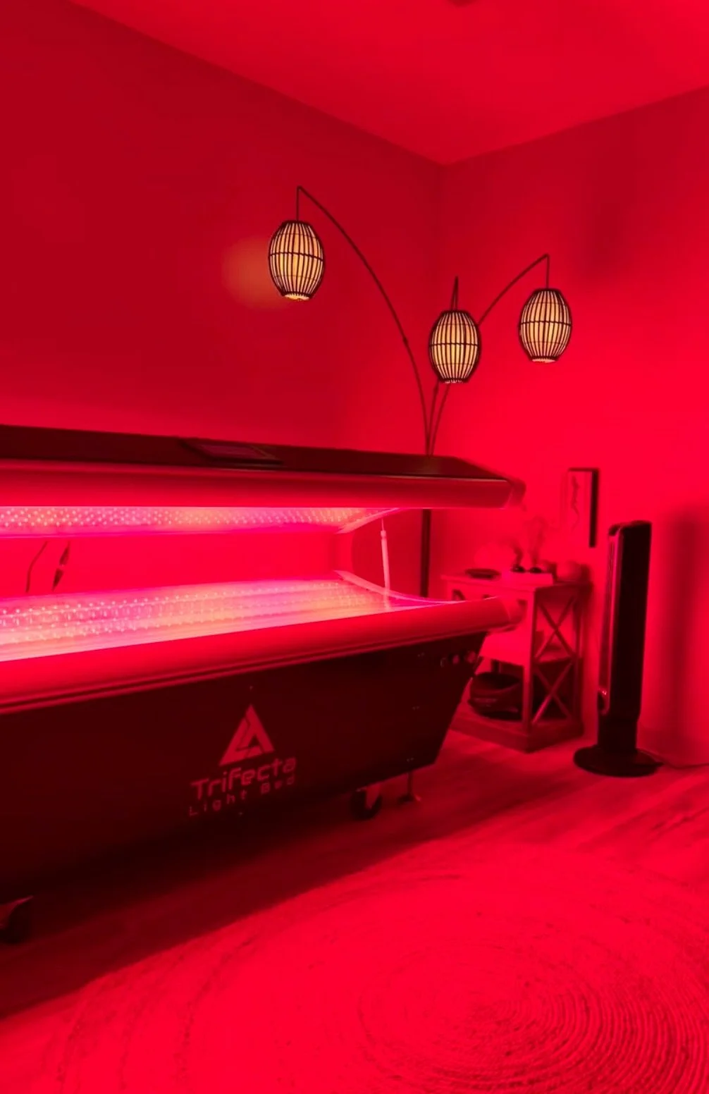 Indoor room with pink lighting, featuring a light therapy machine, a standing fan, a small table, and hanging Asian-style lamps.