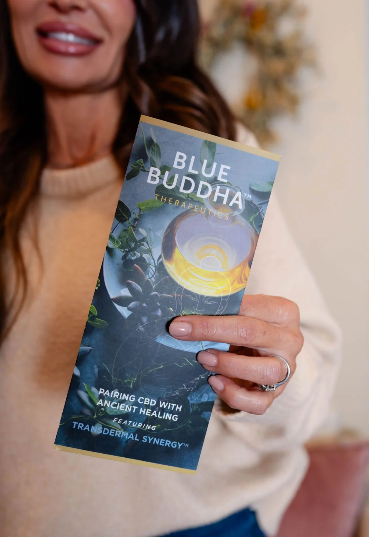 A woman holding a brochure titled 'Blue Buddha Therapeutics' with a graphic of a glass of golden liquid and plants, promoting pairing CBD with ancient healing.