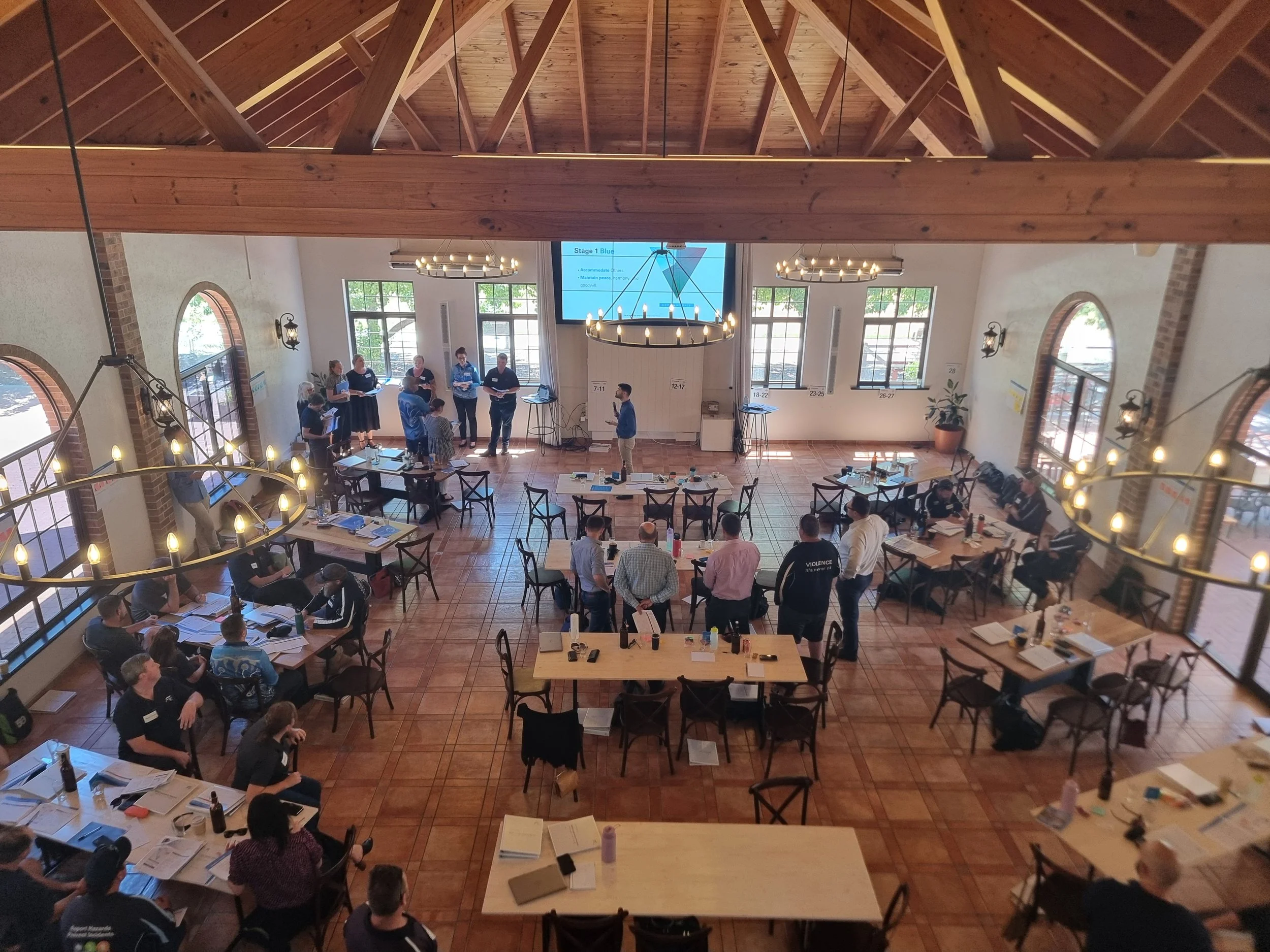 Conference or workshop held in a well-lit, spacious room with chairs, tables, and a large projector screen at the front, with people participating in discussions and presentations.