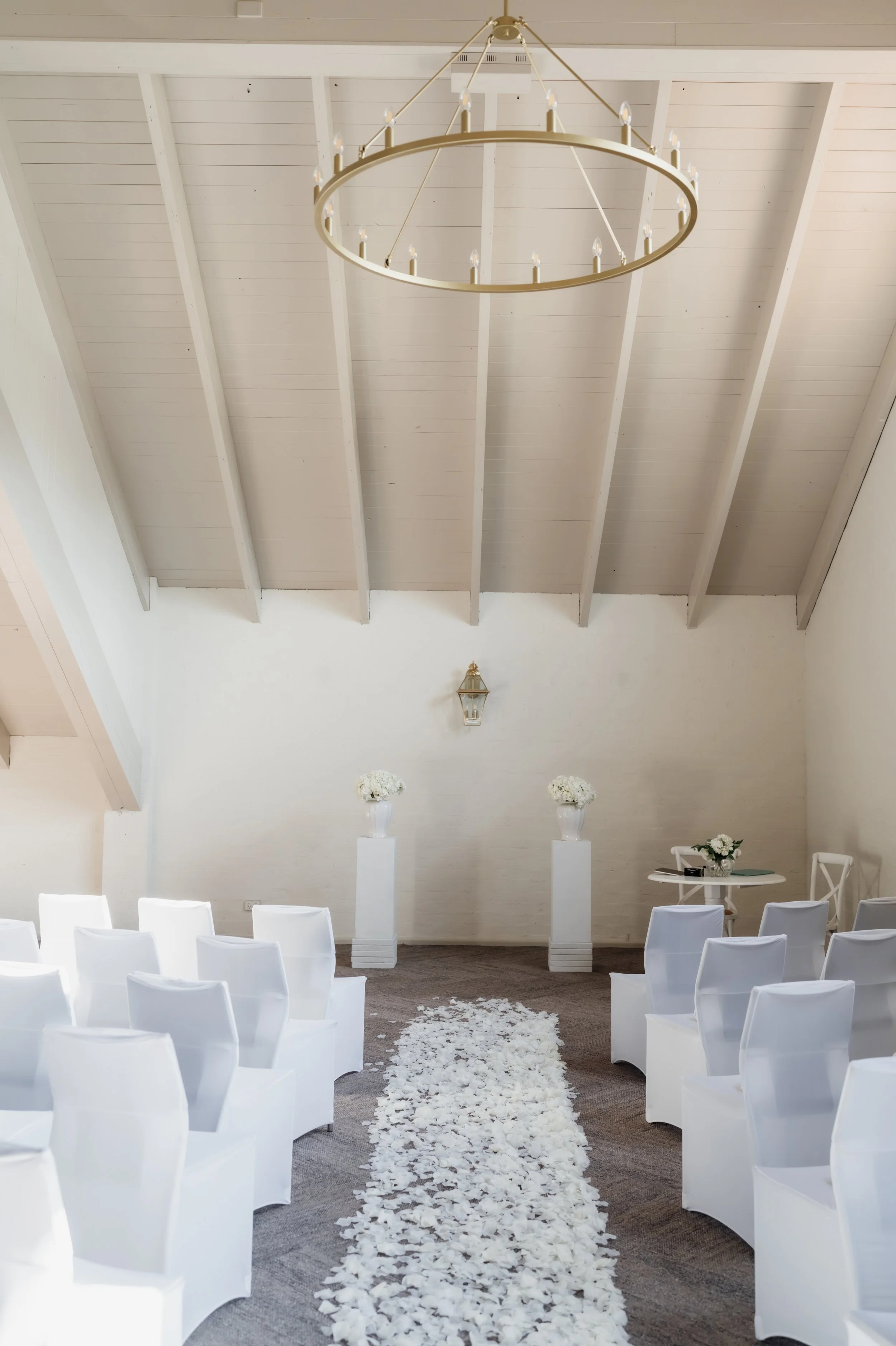 Empty wedding ceremony room with white chairs arranged on either side of a white flower petal aisle, two tall white pedestals with white floral arrangements at the front, a small table with floral decoration to the right, and ceiling with a gold chan