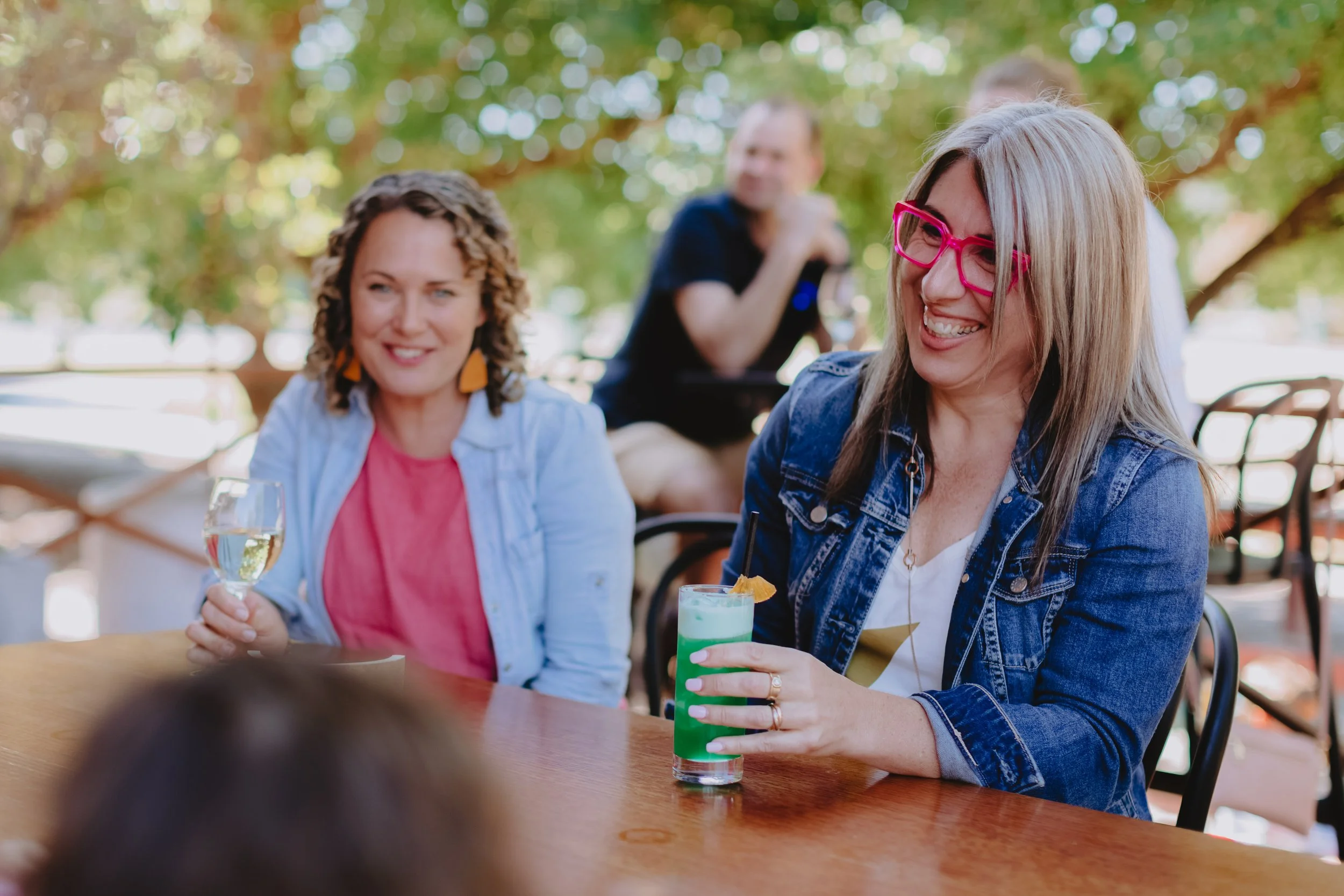 Two women sitting at a wooden table outdoors, one holding a glass of white wine and the other a colorful cocktail, both smiling and enjoying a social gathering under a leafy tree, with a man in the background.